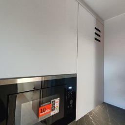 POLI IMAGER - Minimalistyczna kuchnia: białe szafki górne z czarnymi, ukośnymi akcentami wentylacyjnymi i wbudowaną czarną mikrofalówką Whirlpool, na blacie imitującym marmur.