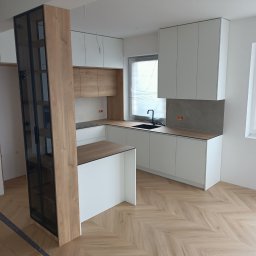 POLI IMAGER - Jasna kuchnia z wyspą i drewnianymi akcentami, parkiet w jodełkę. Okno zasłonięte folią. Widoczna przeszklona witryna. Minimalistyczny design, nowoczesne wykończenie.