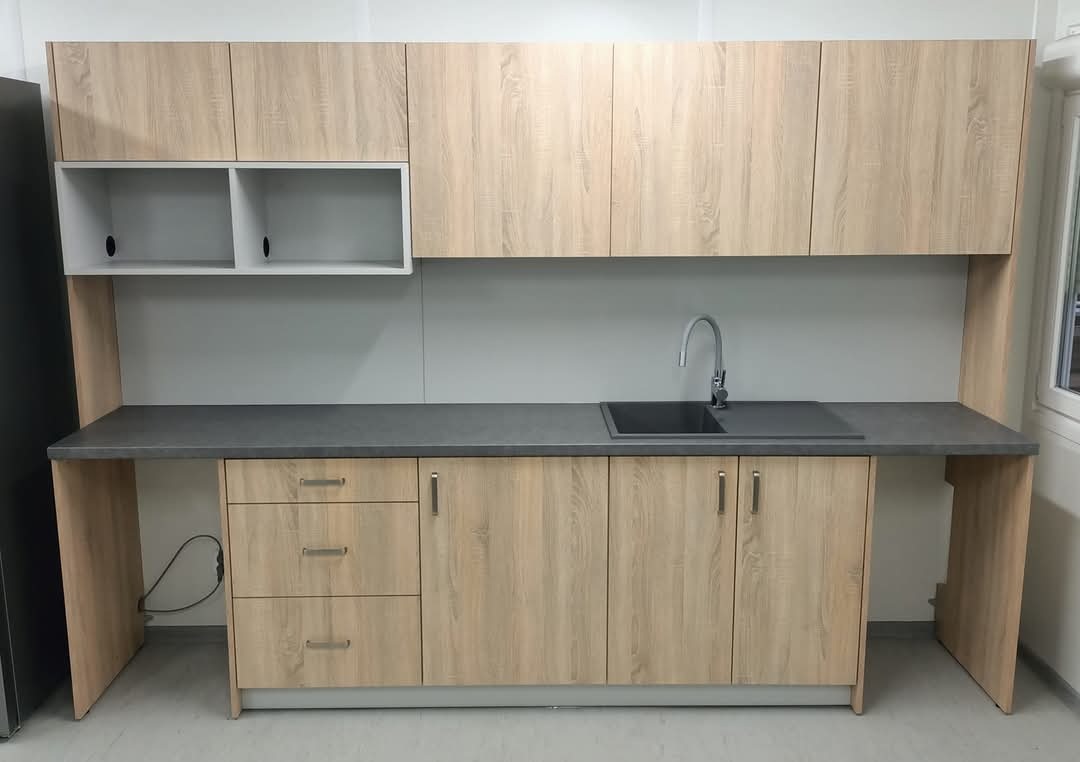 Jasna zabudowa kuchenna z drewnopodobnym frontem, szary blat z wbudowanym zlewozmywakiem i chromowaną baterią, górne szafki z otwartą półką.