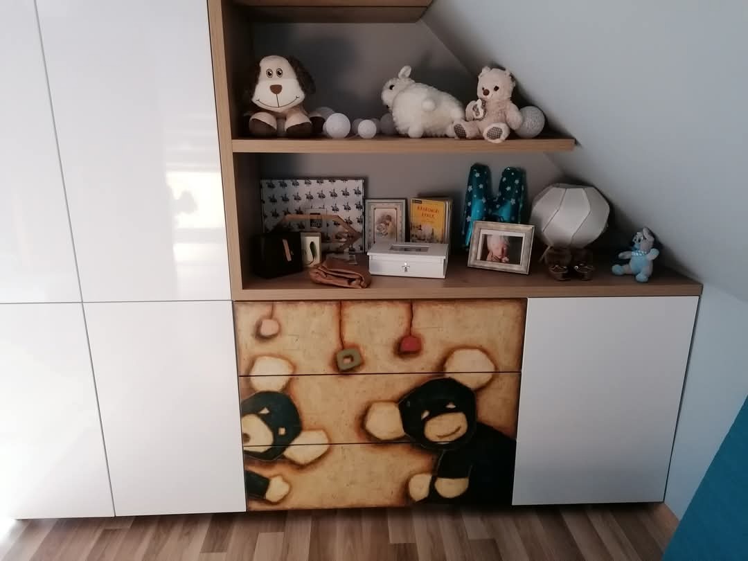 Biała szafa wnękowa z półkami pod skosem, ozdobiona rysunkami niedźwiedzi na frontach szuflad, na półkach pluszaki i dekoracje.