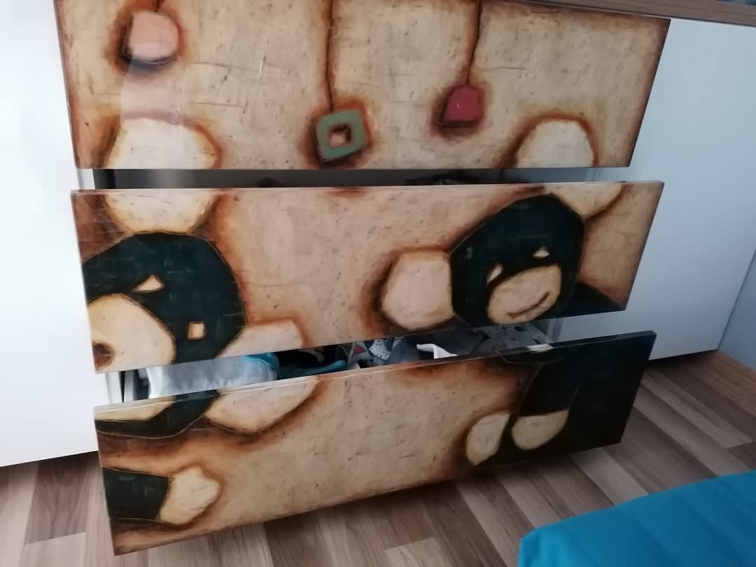 Komoda z trzema szufladami zdobiona artystycznymi rysunkami niedźwiadków i abstrakcyjnych kształtów w ciepłych, beżowo-brązowych tonacjach, na tle białej ściany i drewnianej podłogi.