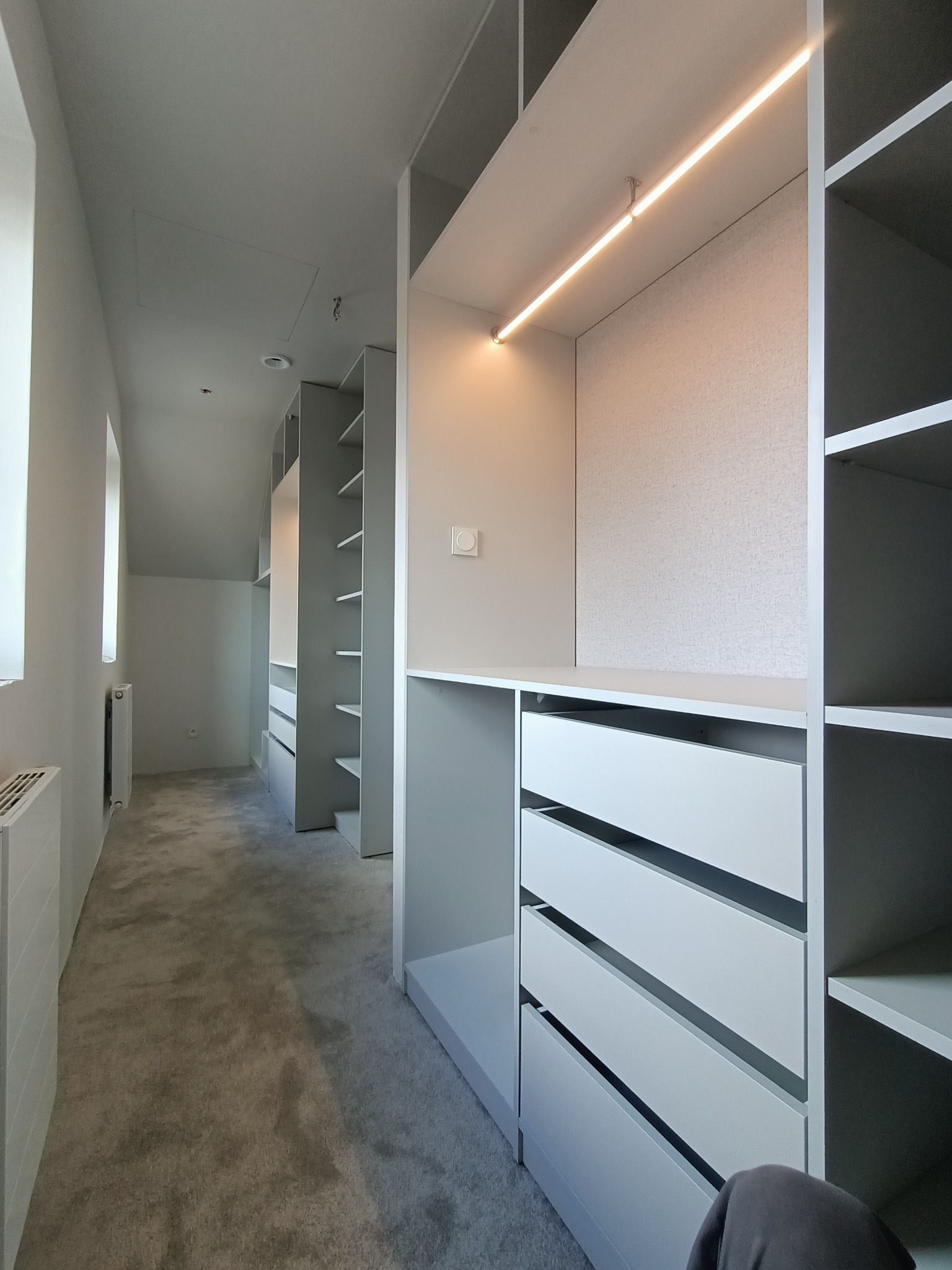 Długa, wąska garderoba w odcieniach szarości z oświetleniem LED podszafkowym, szufladami bez uchwytów i otwartymi półkami, widok na całą długość pomieszczenia.