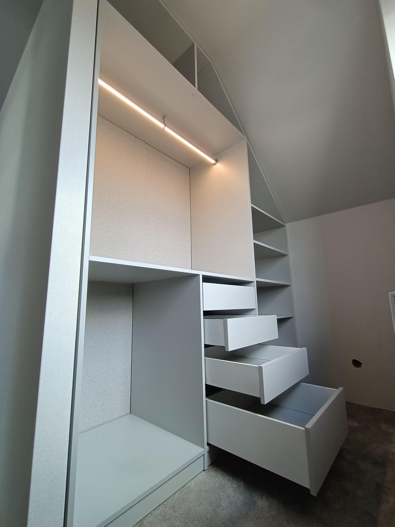 Biała szafa wnękowa z otwartymi szufladami i oświetleniem LED wbudowanym w półki, dopasowana do skosu dachu.
