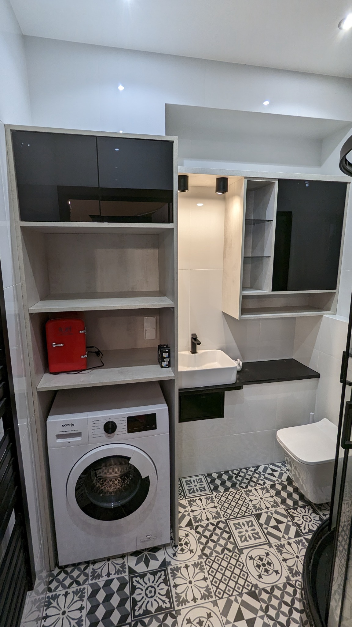 Nowoczesna łazienka z pralką zabudowaną w regale, umywalką nablatową z czarną armaturą, szafkami wiszącymi z czarnymi frontami i wzorzystą podłogą.