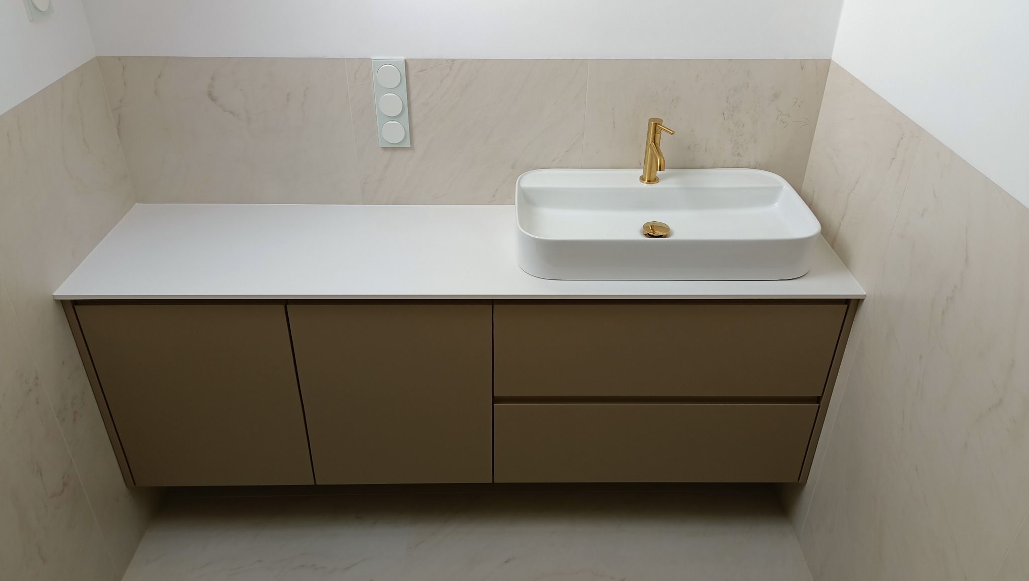 Nowoczesna łazienka z beżową szafką pod umywalką, białą prostokątną umywalką nablatową i złotą baterią. Na ścianach płytki imitujące marmur. Widoczny włącznik światła z trzema okrągłymi przyciskami.