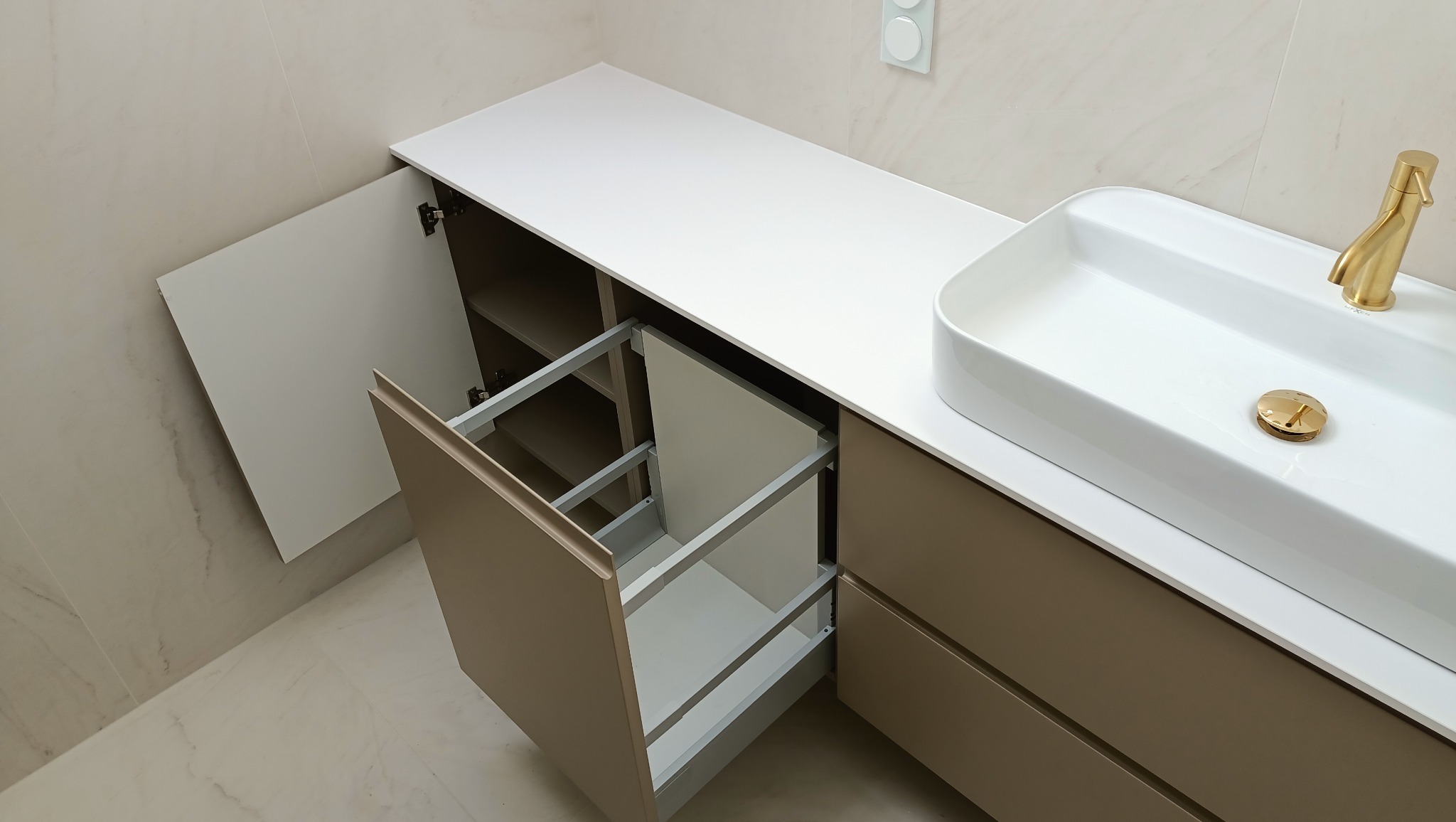 Nowoczesna szafka łazienkowa z beżowymi frontami, białym blatem i umywalką nablatową. Otwarta szafka z wysuwanym koszem cargo i półkami.