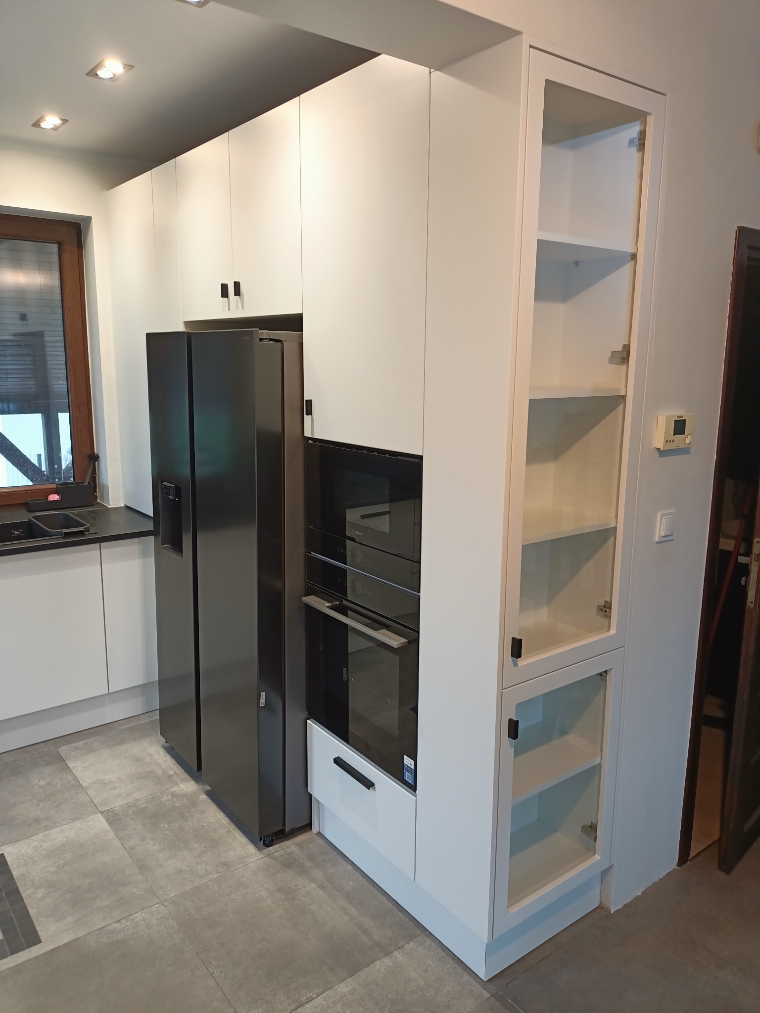 Nowoczesna, biała zabudowa kuchenna z czarnym sprzętem AGD: lodówka side-by-side, piekarnik i szklana witryna. Minimalistyczne uchwyty. Szare płytki podłogowe.