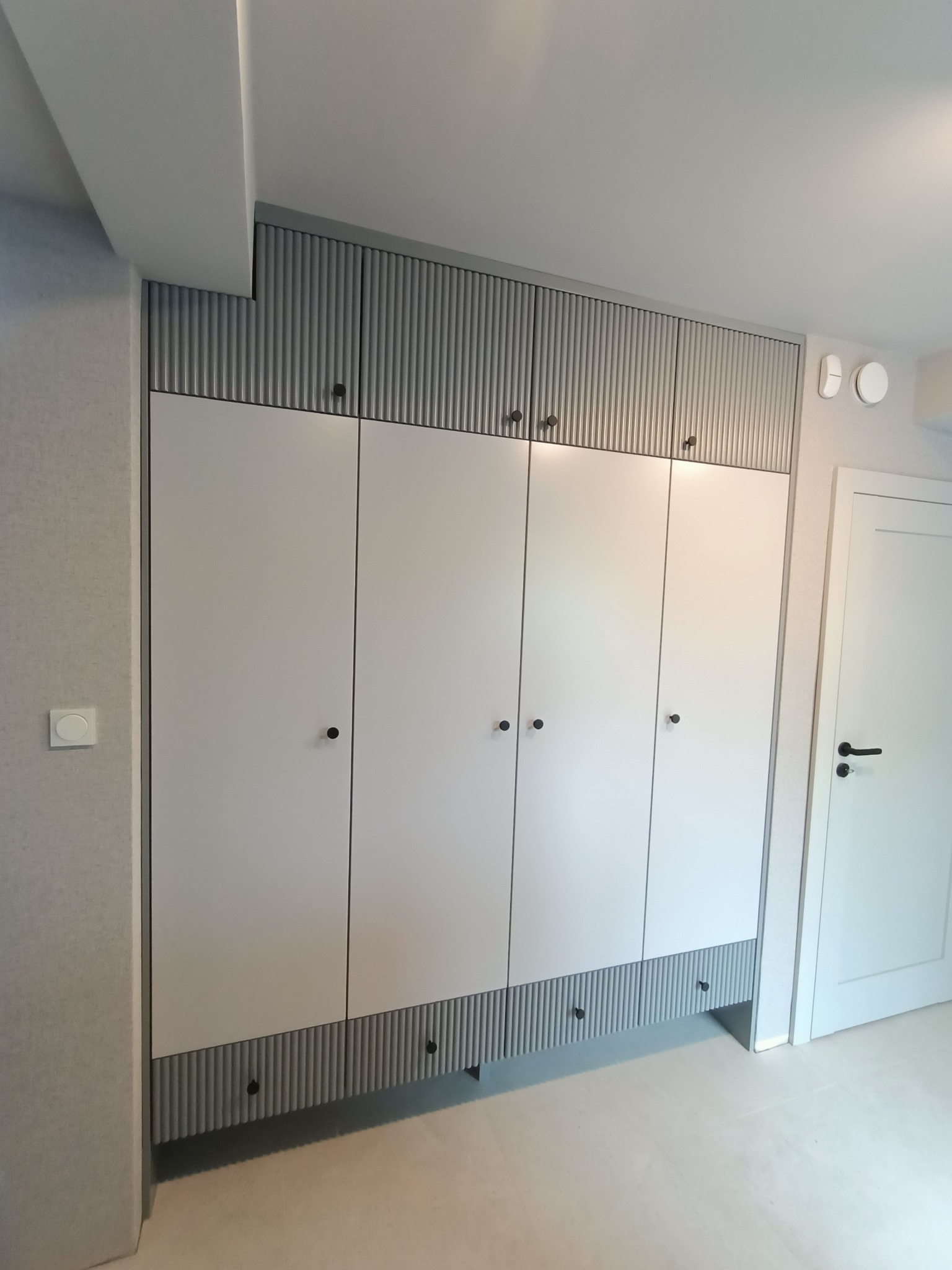 Biała szafa wnękowa z ryflowanymi elementami dekoracyjnymi, czarne okrągłe uchwyty, dopasowana kolorystycznie do ścian i drzwi w nowoczesnym wnętrzu.