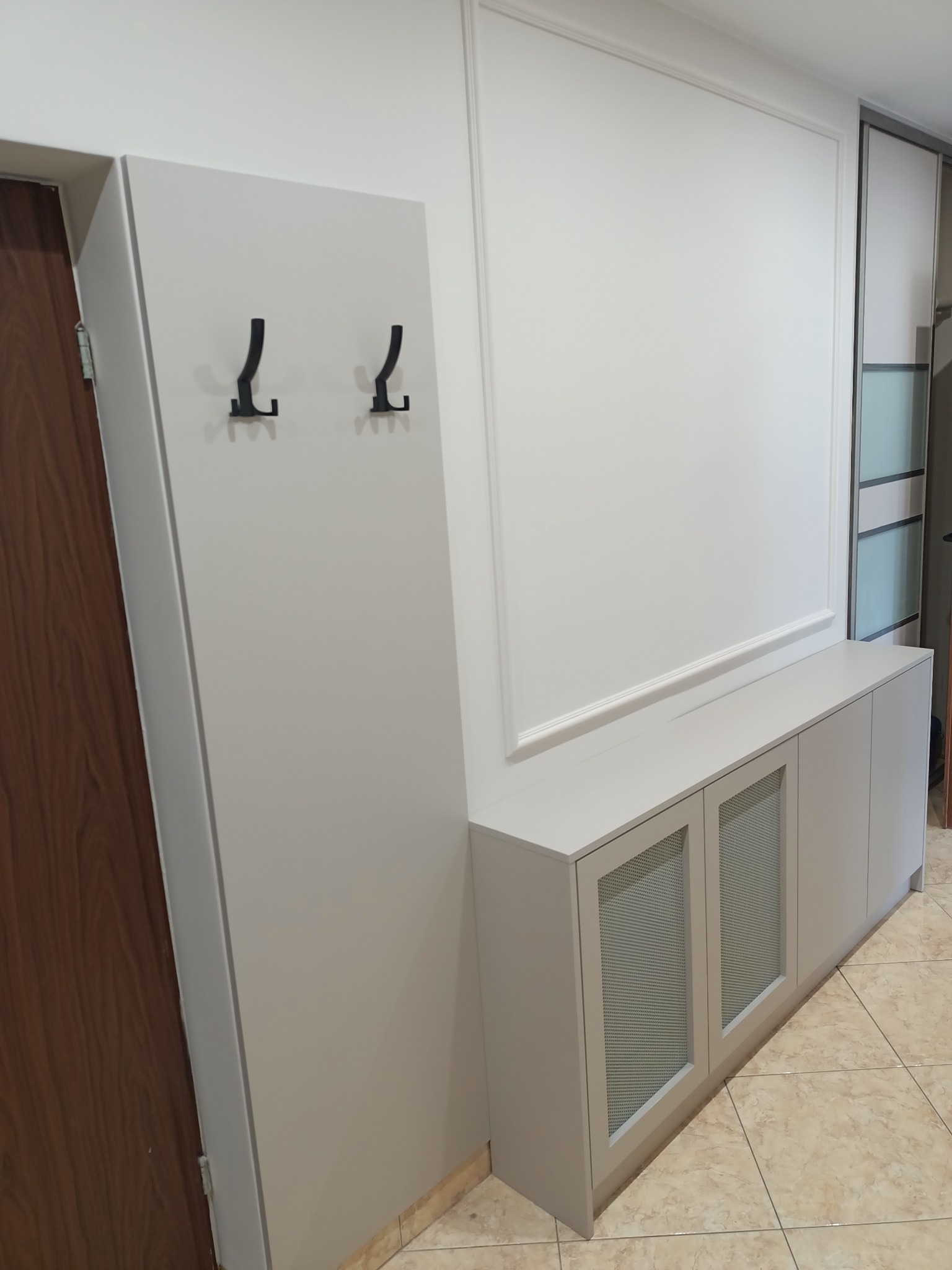 Hall zabudowany szafką z ażurowymi drzwiczkami i panelem wieszakowym z haczykami, całość w jasnych odcieniach, z widocznymi detalami wykończenia.