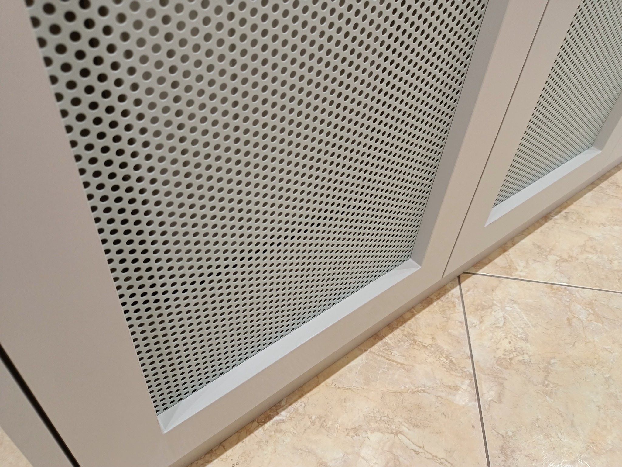 Szara, perforowana szafka wnękowa z widoczną podłogą z płytek imitujących marmur. Detal wykończenia wnętrza, bliski kadr.