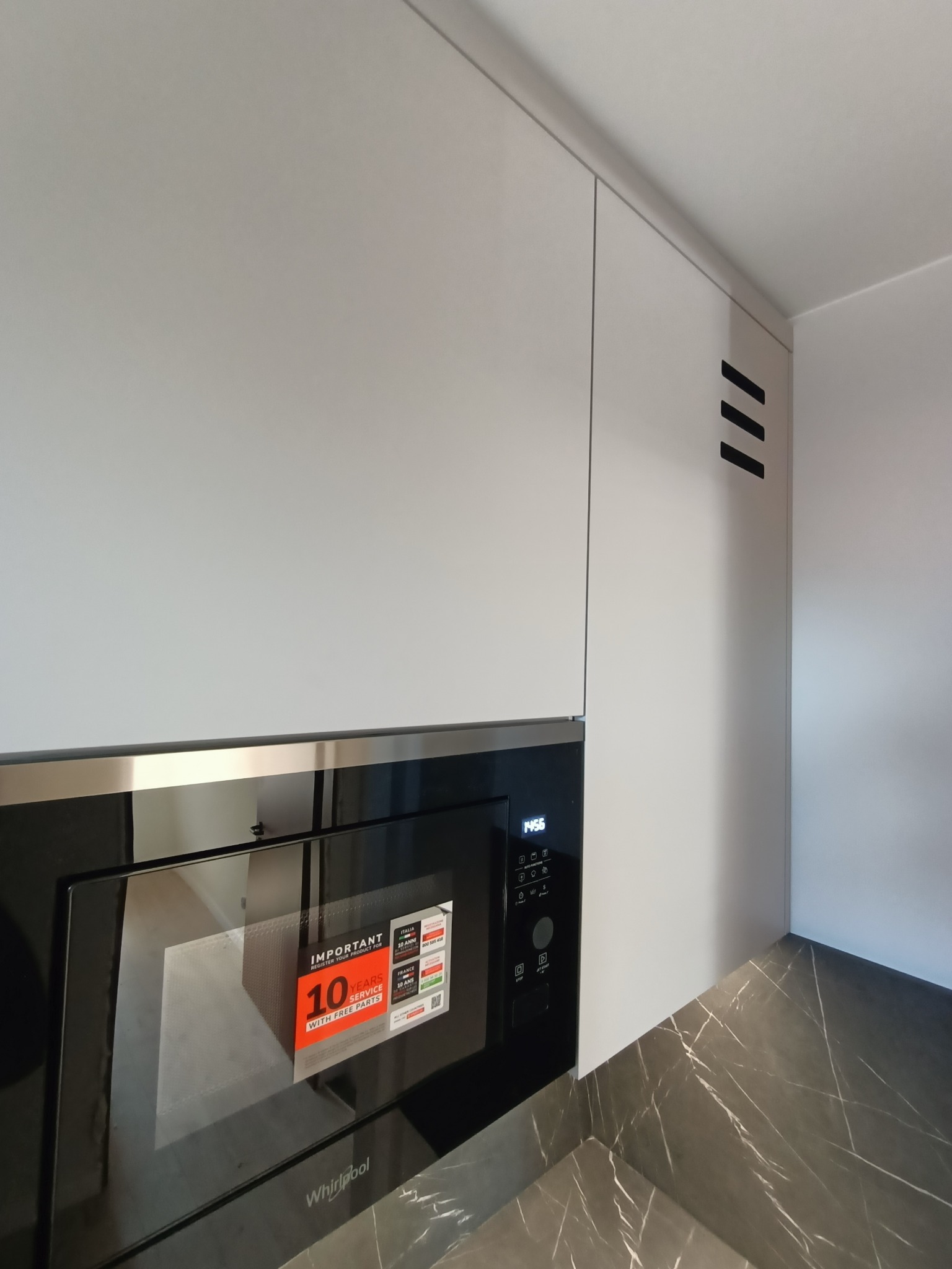 Minimalistyczna kuchnia: białe szafki górne z czarnymi, ukośnymi akcentami wentylacyjnymi i wbudowaną czarną mikrofalówką Whirlpool, na blacie imitującym marmur.