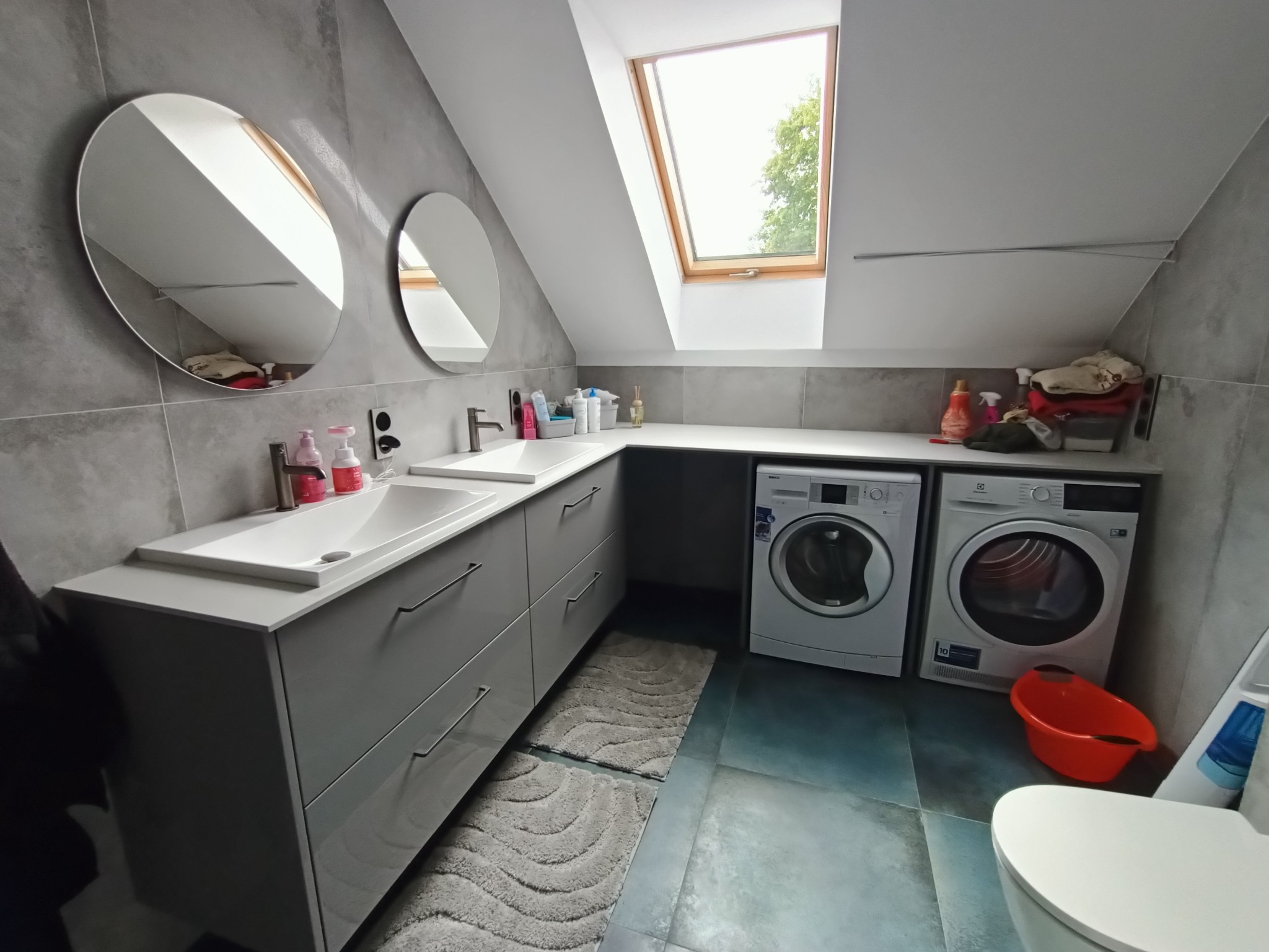 Szara łazienka na poddaszu ze skośnym oknem dachowym. Dwie okrągłe umywalki, pralka i suszarka. Minimalistyczny design z szarymi płytkami i dywanikami.