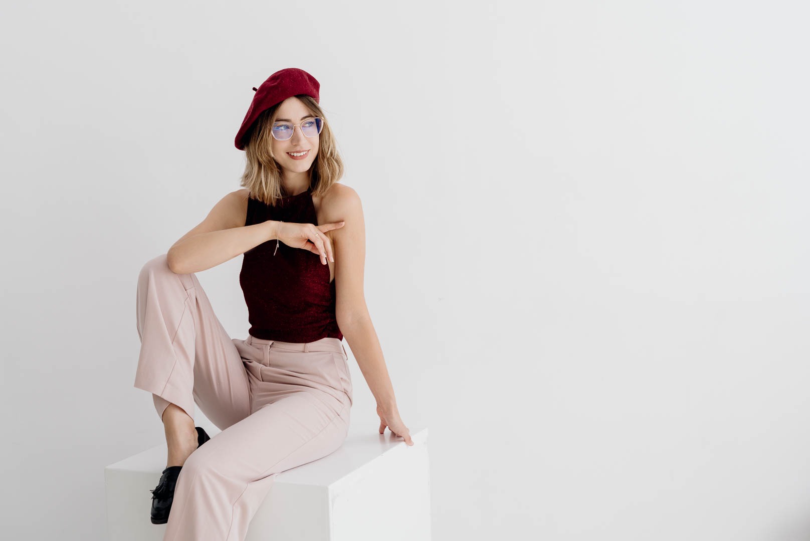 Uśmiechnięta kobieta w bordowym berecie i okularach, siedząca na białym sześcianie, wskazuje palcem w prawą stronę w jasnym studiu.