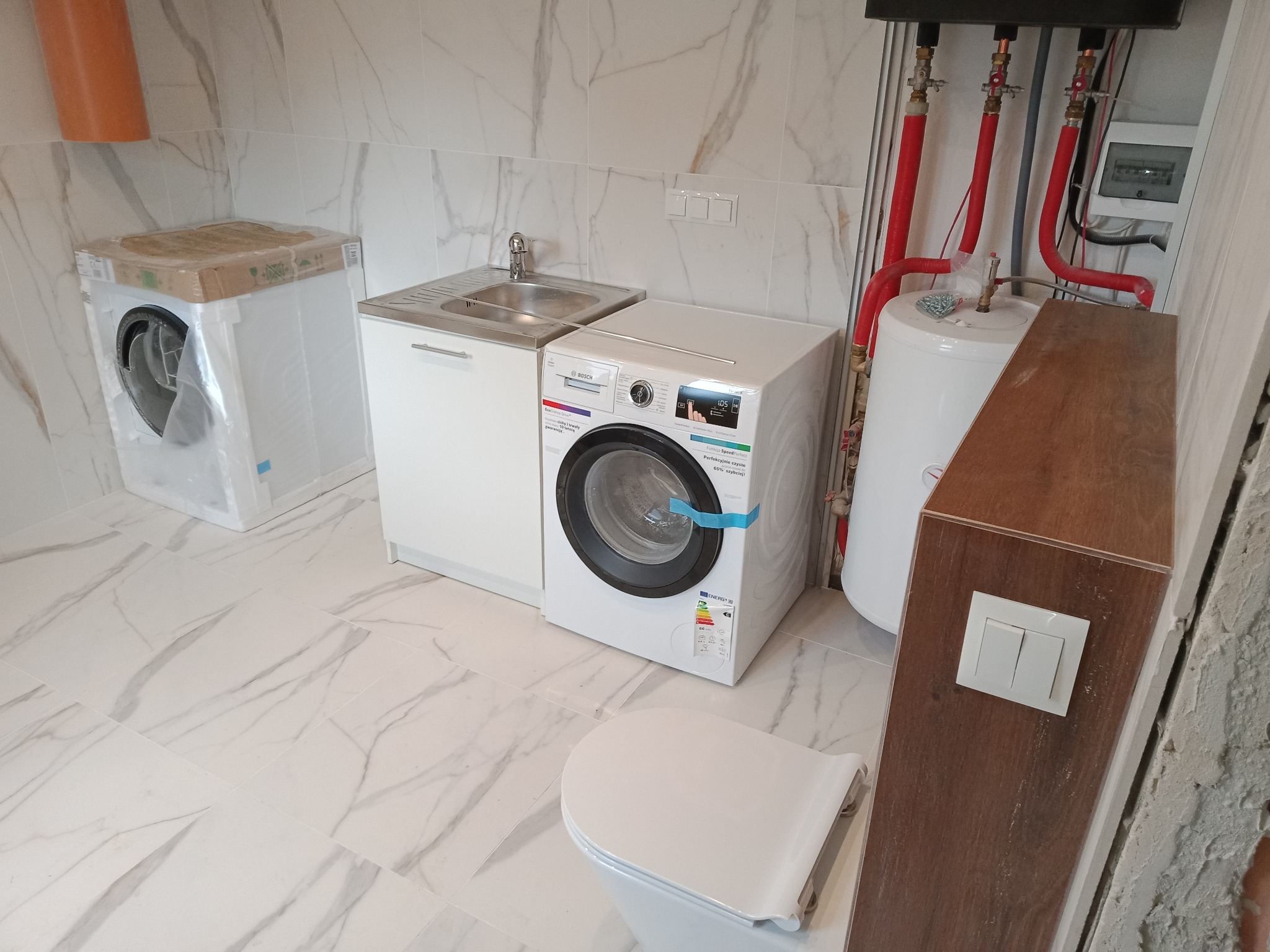 Nowa pralka w folii ochronnej obok umywalki z szafką, białe marmurowe płytki na ścianach i podłodze, bojler z czerwonymi rurami, widok na fragment toalety i włącznik światła na drewnianej obudowie.