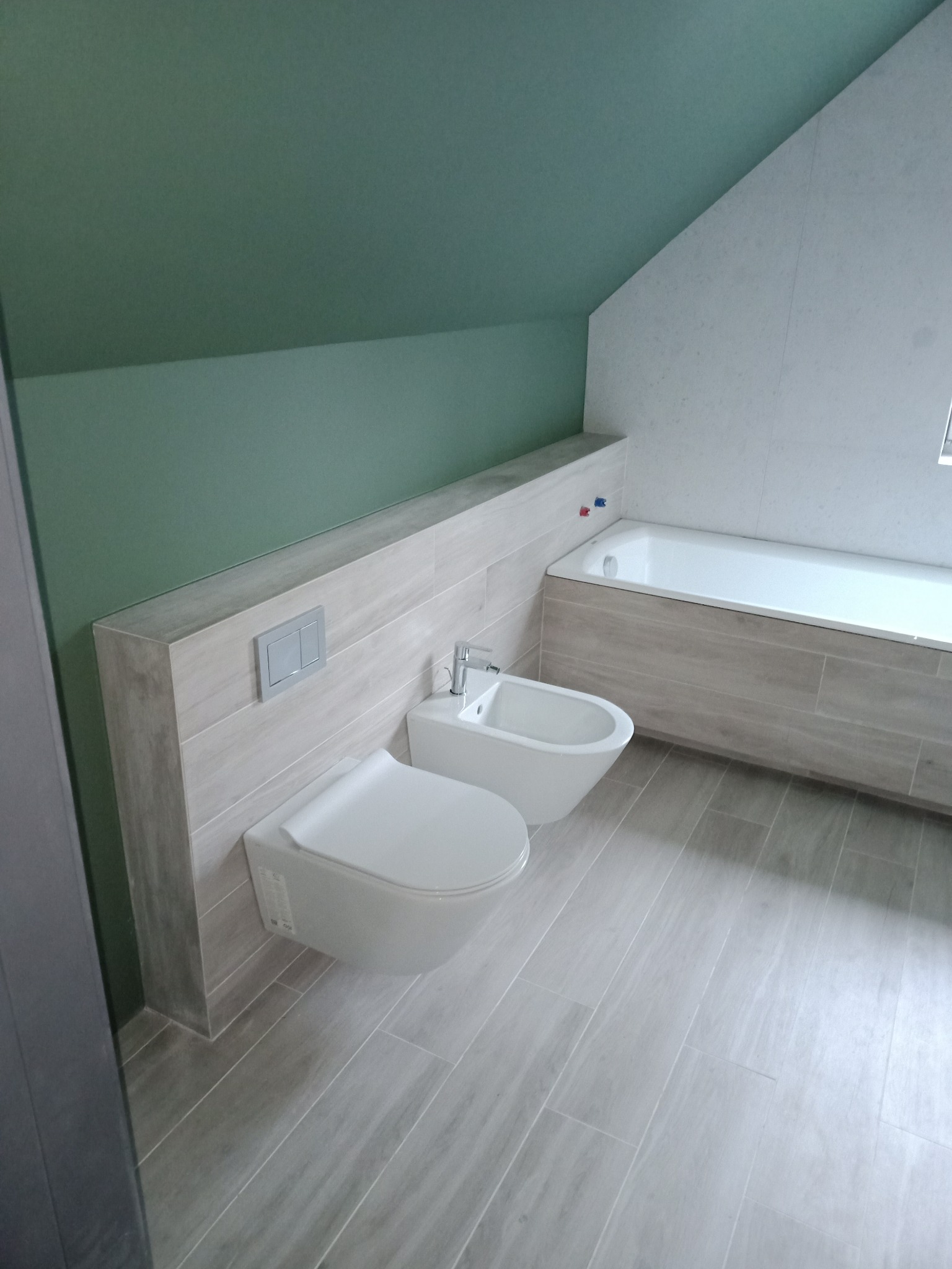 Nowoczesna łazienka na poddaszu z wanną, toaletą i bidetem w jasnych płytkach imitujących drewno, zielony sufit pod skosem.