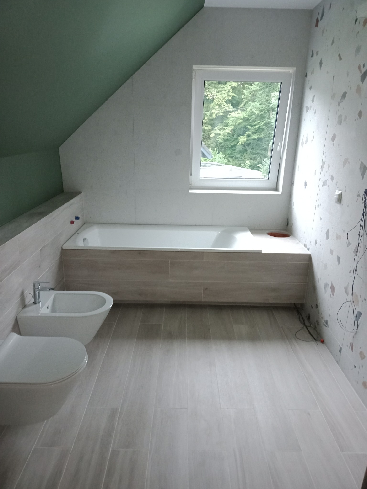 Wykończona łazienka na poddaszu ze skośnym sufitem, z białą wanną obudowaną płytkami imitującymi drewno, białą armaturą sanitarną, płytkami podłogowymi imitującymi drewno i ścianą z mozaiką, widok...