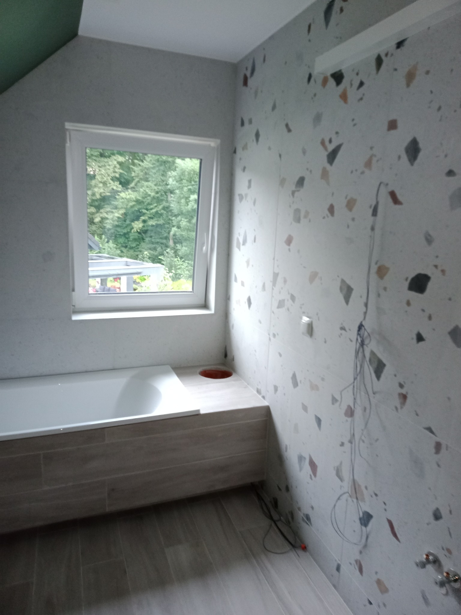 Łazienka w trakcie remontu w Mazańcowicach, widoczna wanna obudowana płytkami imitującymi drewno, ściany wyłożone płytkami w stylu lastryko, okno z widokiem na zieleń, wystające kable...