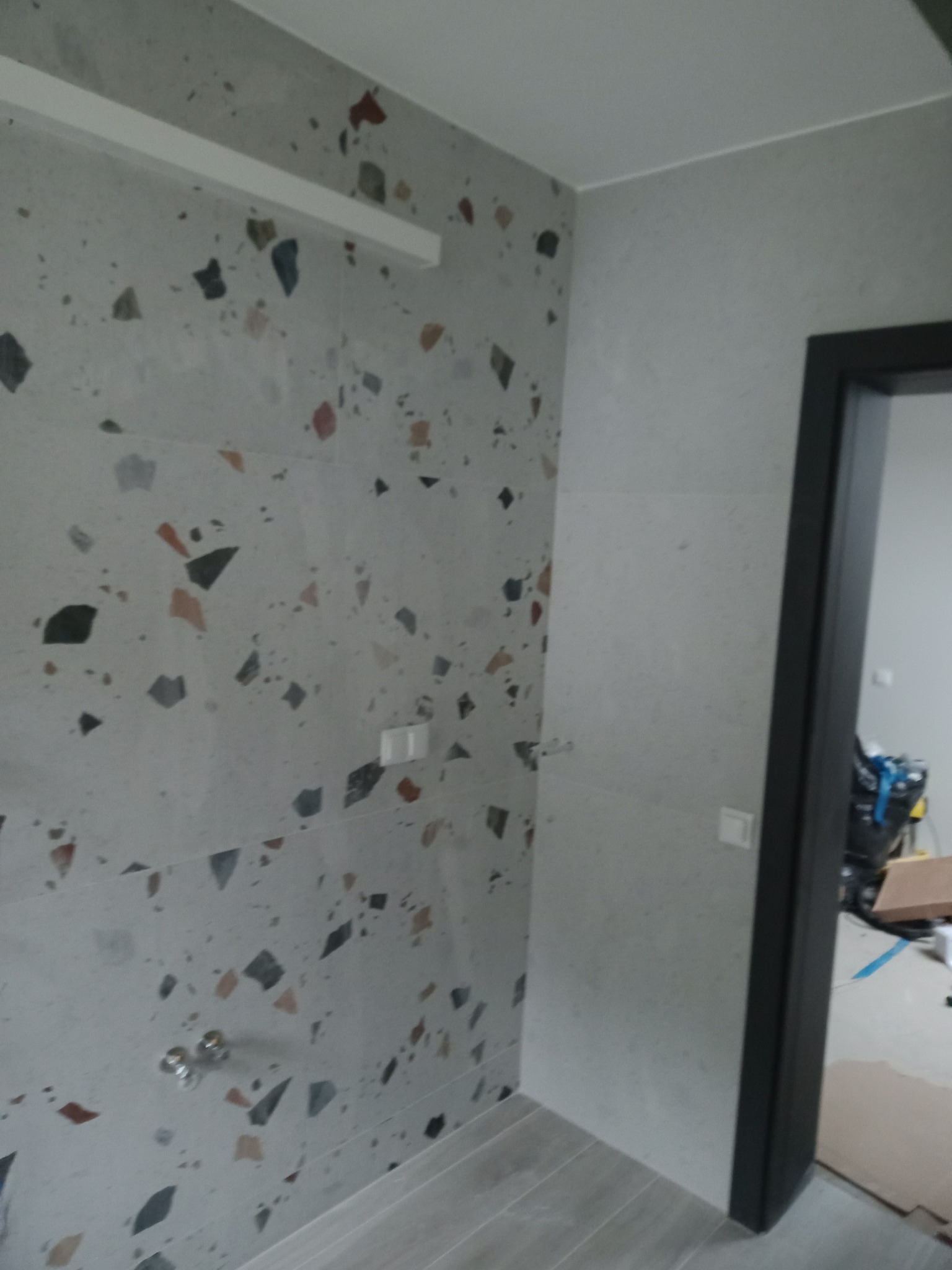 Ściana wyłożona płytkami imitującymi lastryko w łazience, widoczne chromowane zawory, biały włącznik światła i fragment czarnej ościeżnicy drzwiowej.