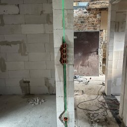 Dawid Piskorz Usługi Elektryczne - Ściana z pustaków z zamontowanymi puszkami elektrycznymi, pion wyznaczony laserowo. Widoczne przewody i narzędzia w surowym wnętrzu budynku w trakcie prac wykończeniowych.