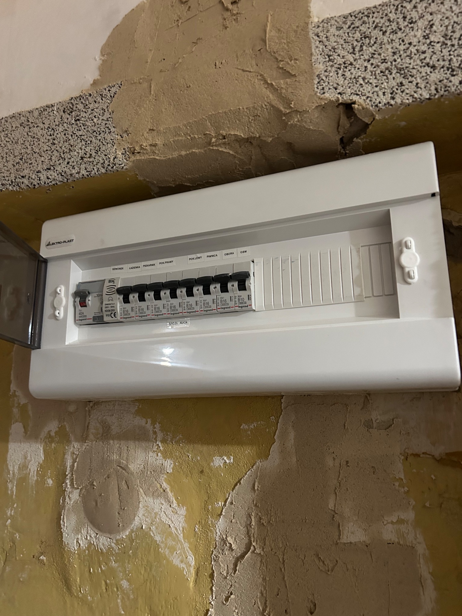 Biała skrzynka elektryczna z bezpiecznikami Legrand zamontowana na żółtej ścianie w trakcie remontu. Widoczna otwarta klapka i opis bezpieczników.