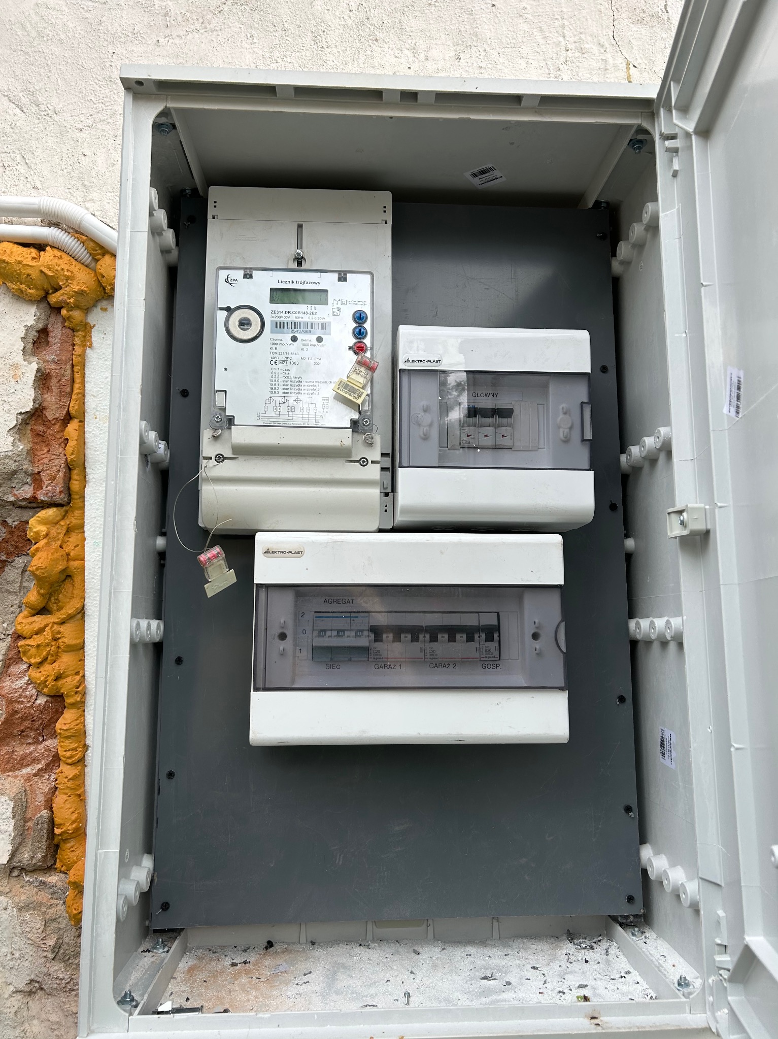 Otwarta skrzynka z licznikiem elektrycznym i bezpiecznikami na szarej płycie montażowej, widoczne przewody i pianka montażowa przy murze.