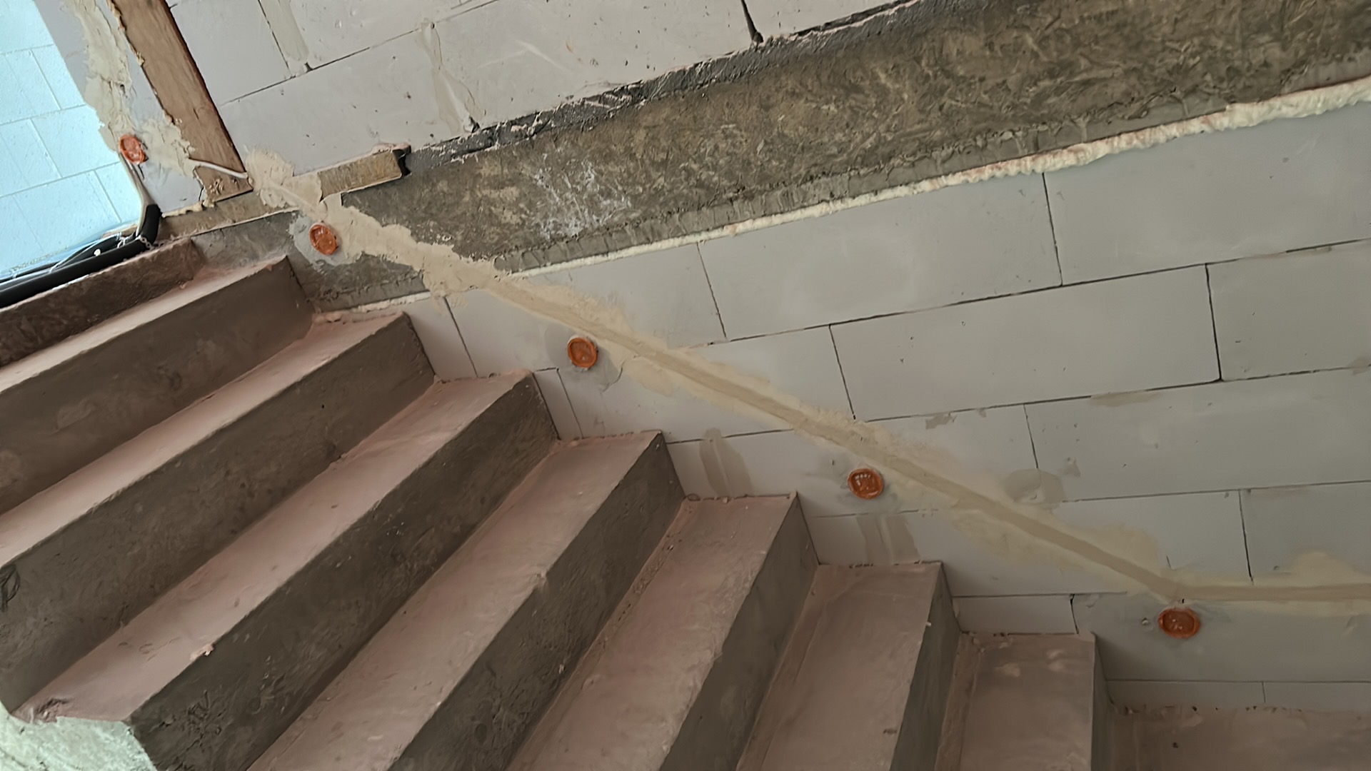 Betonowe schody z instalacją elektryczną w ścianie z bloczków, widoczne pomarańczowe puszki pod gniazdka. Prace w toku, surowy stan wykończenia.