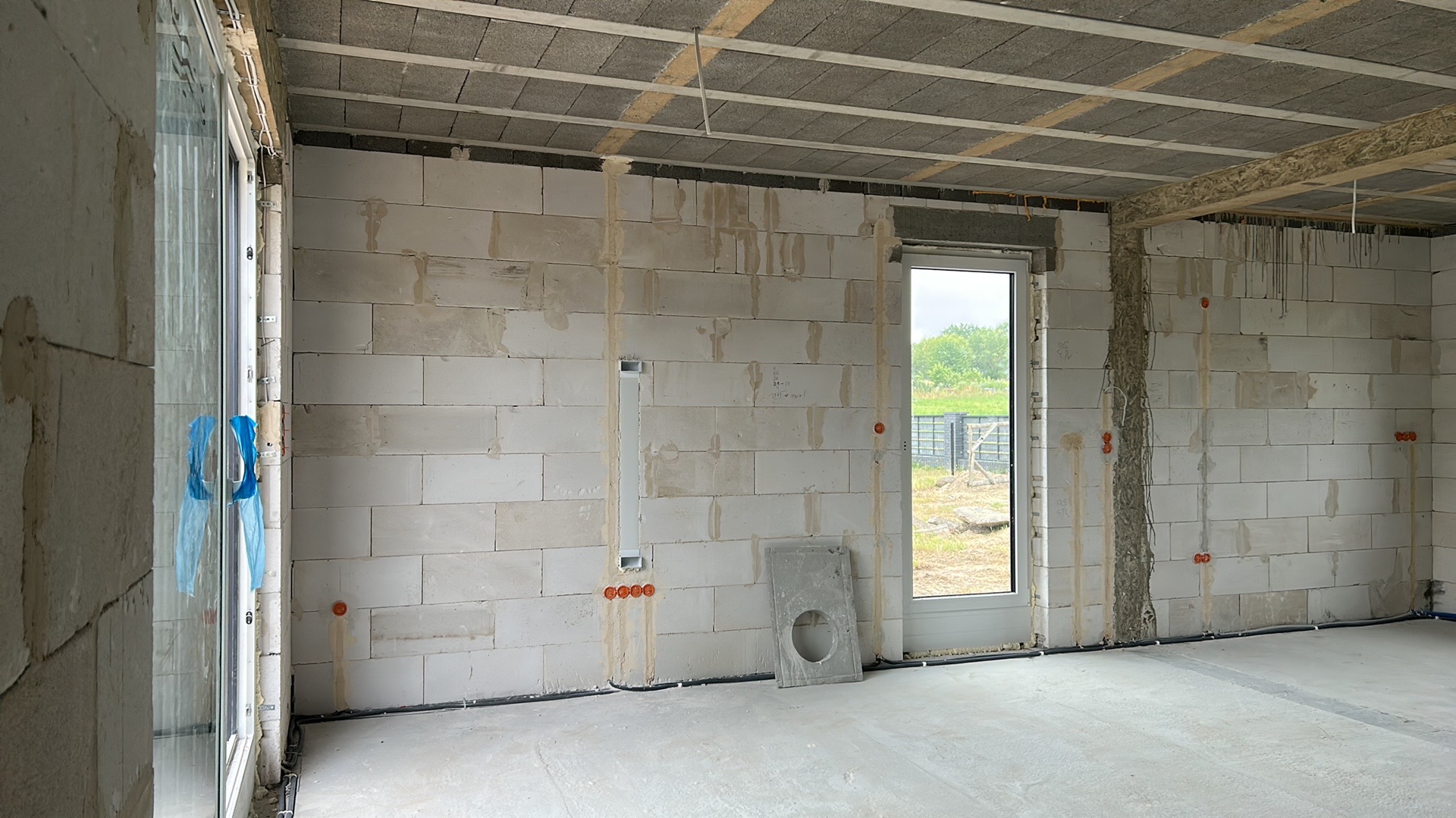 Wnętrze w stanie surowym: ściany z betonu komórkowego z widocznymi puszkami instalacyjnymi i przewodami elektrycznymi. Okno z widokiem na zieleń.