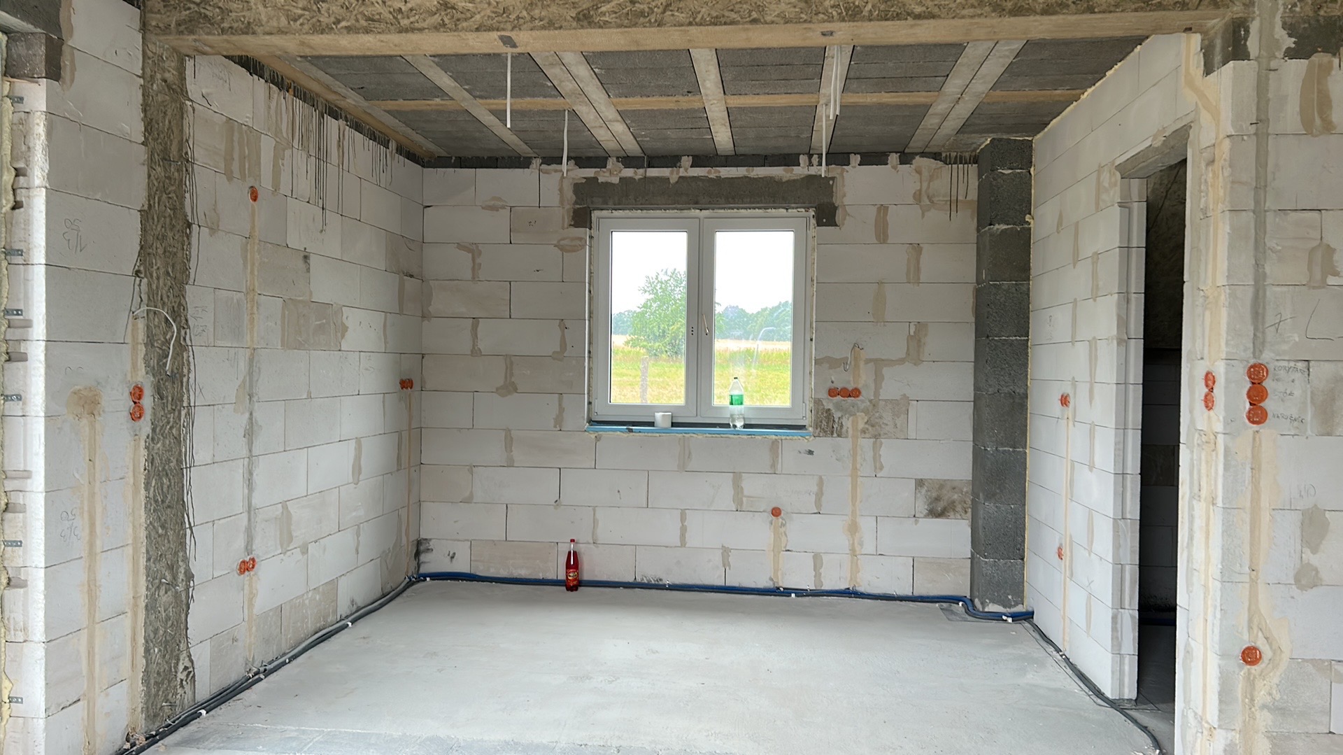 Surowy stan wnętrza z instalacją elektryczną w ścianach z betonu komórkowego. Widoczne puszki instalacyjne i przewody. W tle okno z widokiem na zieleń.