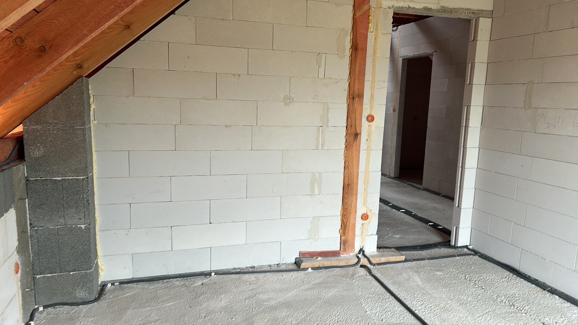 Surowy stan poddasza z instalacją elektryczną w peszlach przy podłodze. Widoczne przewody prowadzone wzdłuż ścian i drewniane elementy konstrukcyjne.