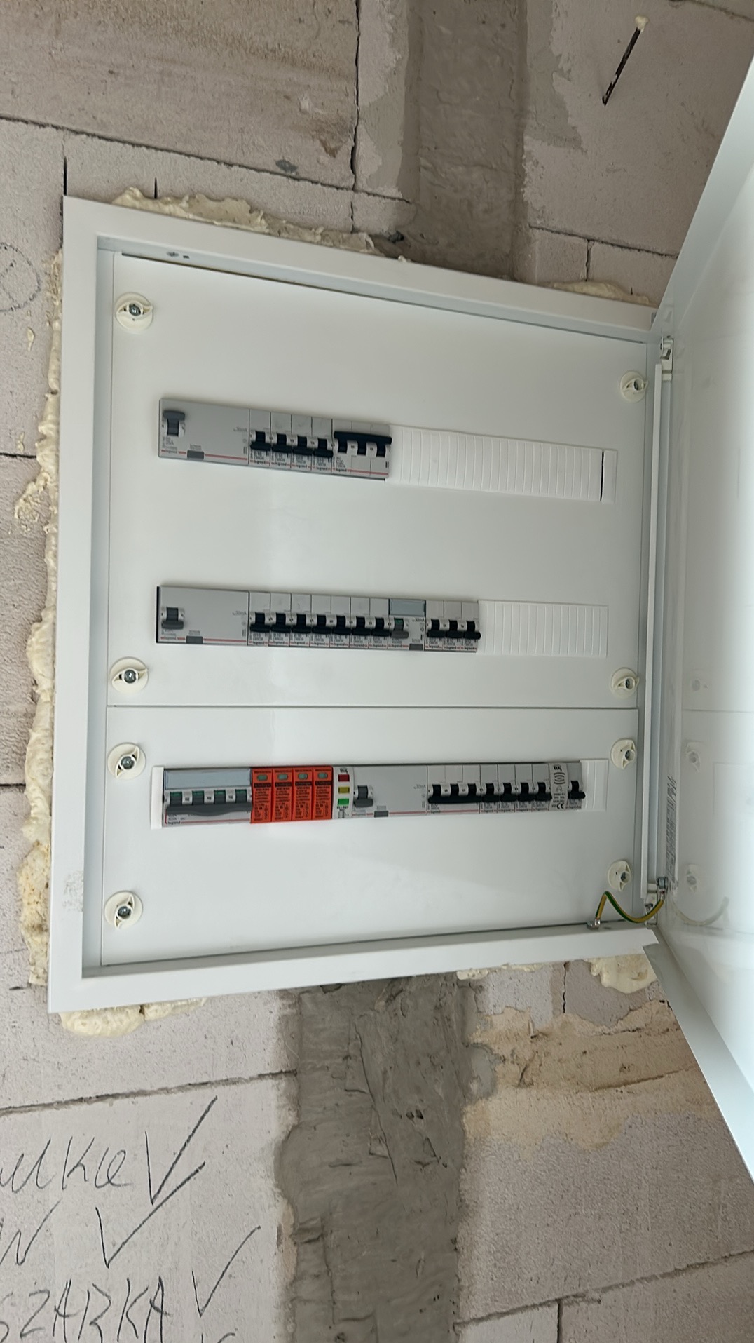 Nowa, otwarta skrzynka rozdzielcza z bezpiecznikami na ścianie z betonu komórkowego, zabezpieczona pianką montażową. Widoczny przewód uziemiający.