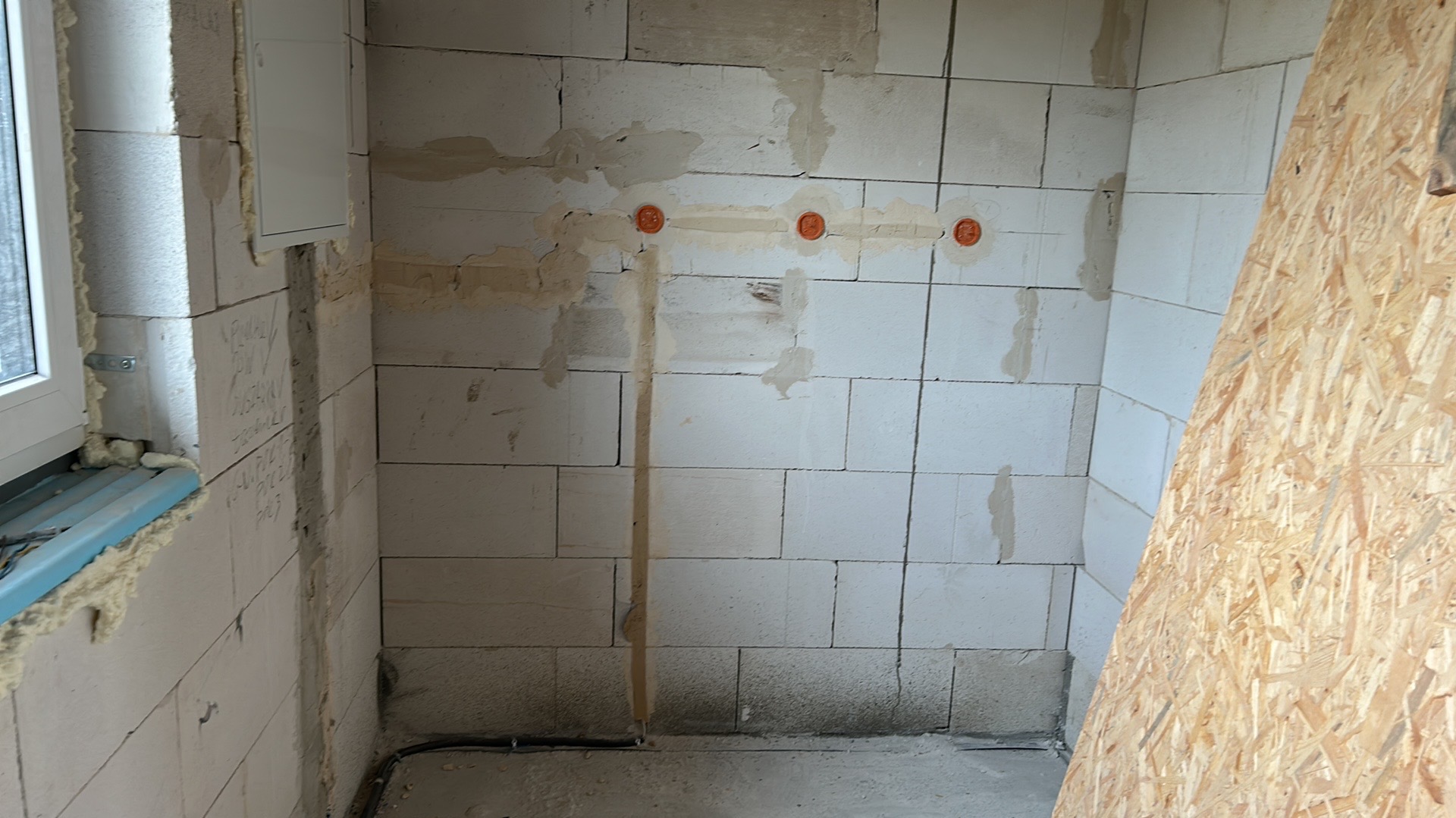 Wnętrze w trakcie prac: ściany z betonu komórkowego z widocznymi puszkami instalacyjnymi w rzędzie oraz bruzdą pionową, czarne kable przy podłodze, fragment okna i płyta OSB.