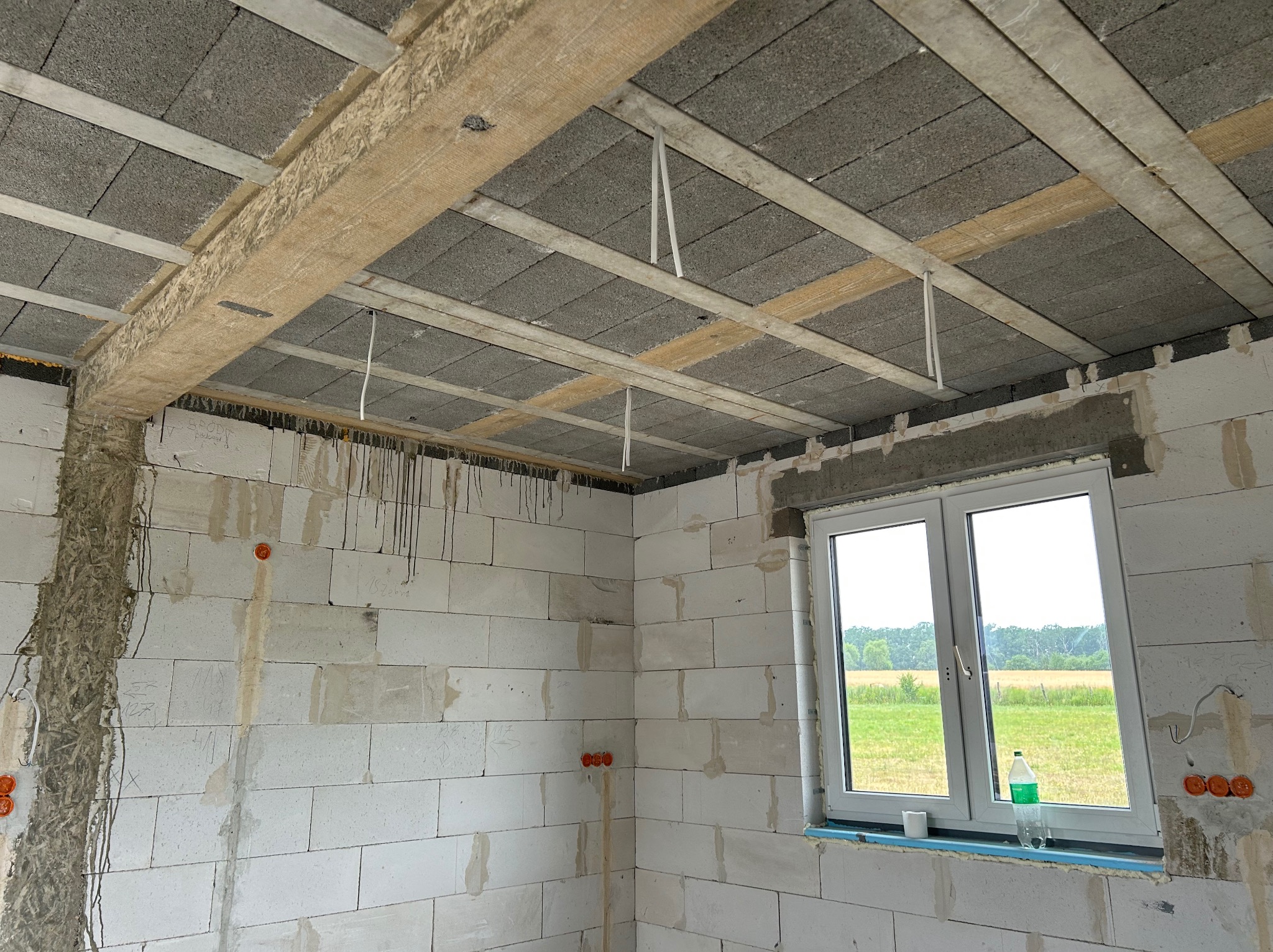 Surowy stan wnętrza w Krośnicach: ściany z białych bloczków, drewniane belki stropowe z wystającymi przewodami. Widoczne puszki instalacyjne.
