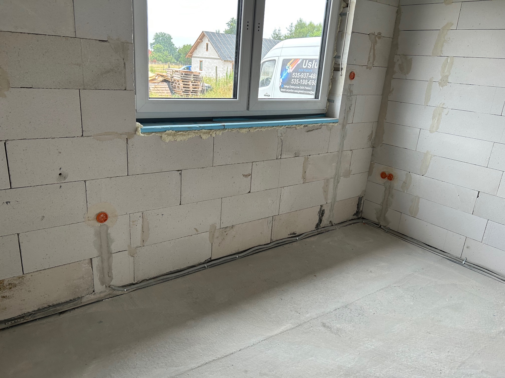 Surowy stan: wnętrze z białymi bloczkami, okno z widokiem na budynek i auto firmy elektrycznej, widoczne kable i puszki elektryczne w ścianach.