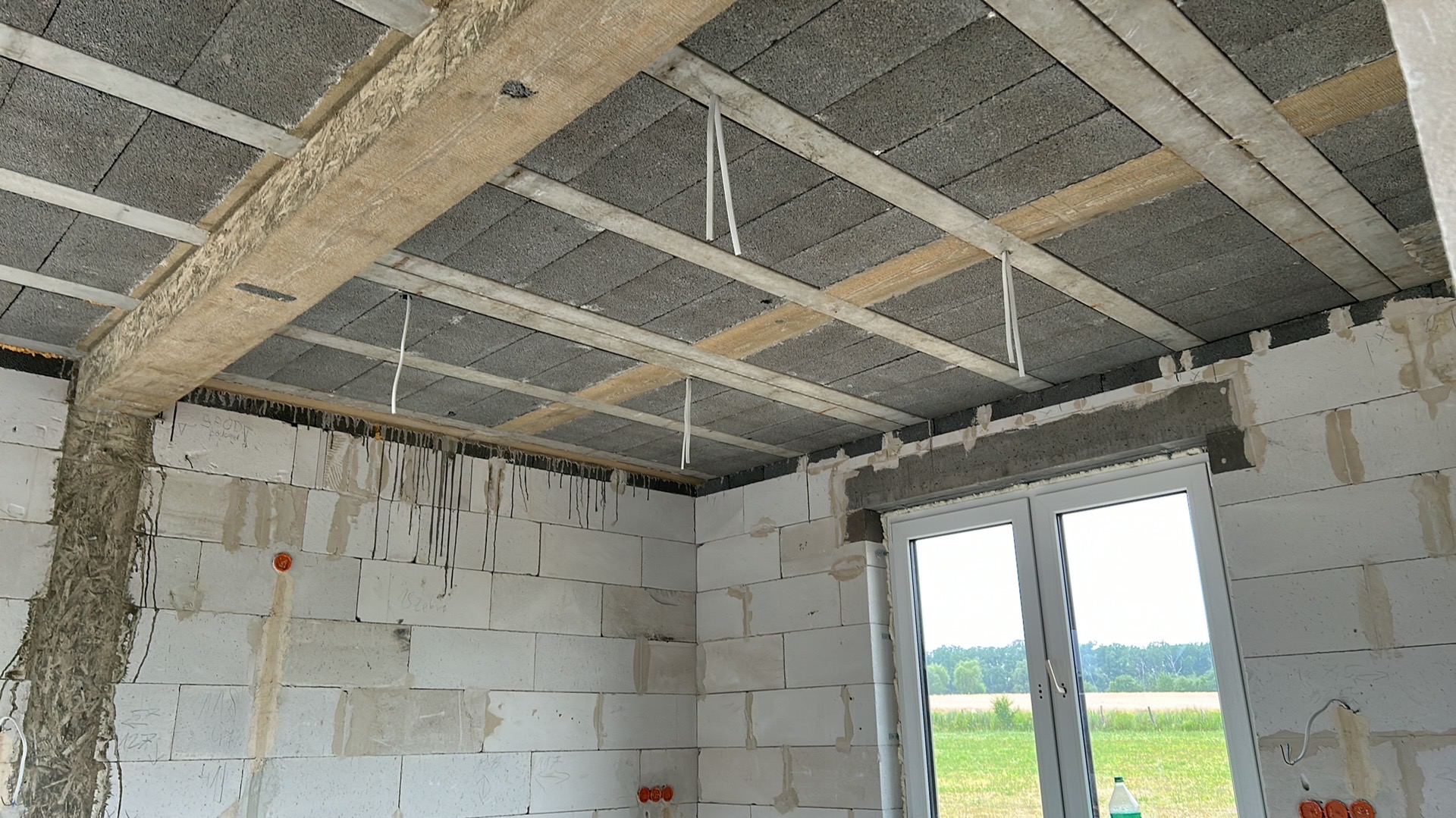 Wnętrze w trakcie budowy: sufit z betonowych bloków, drewniane belki i wystające przewody elektryczne. Widoczne puszki elektryczne w ścianie z białych bloczków.