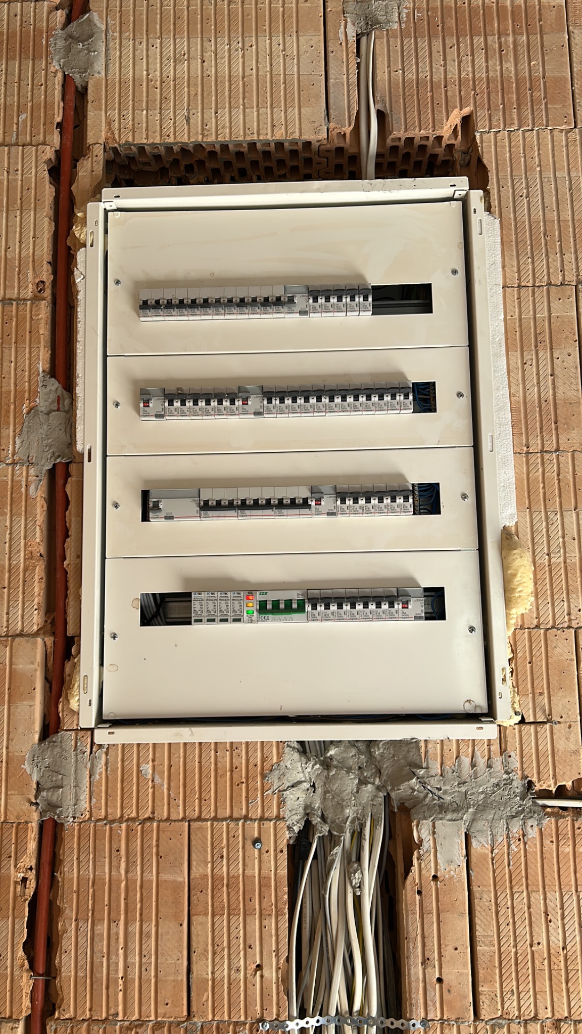 Rozdzielnia elektryczna zamontowana w ścianie z cegły dziurawki, widoczne kable i zabezpieczenia. Instalacja w trakcie prac wykończeniowych.