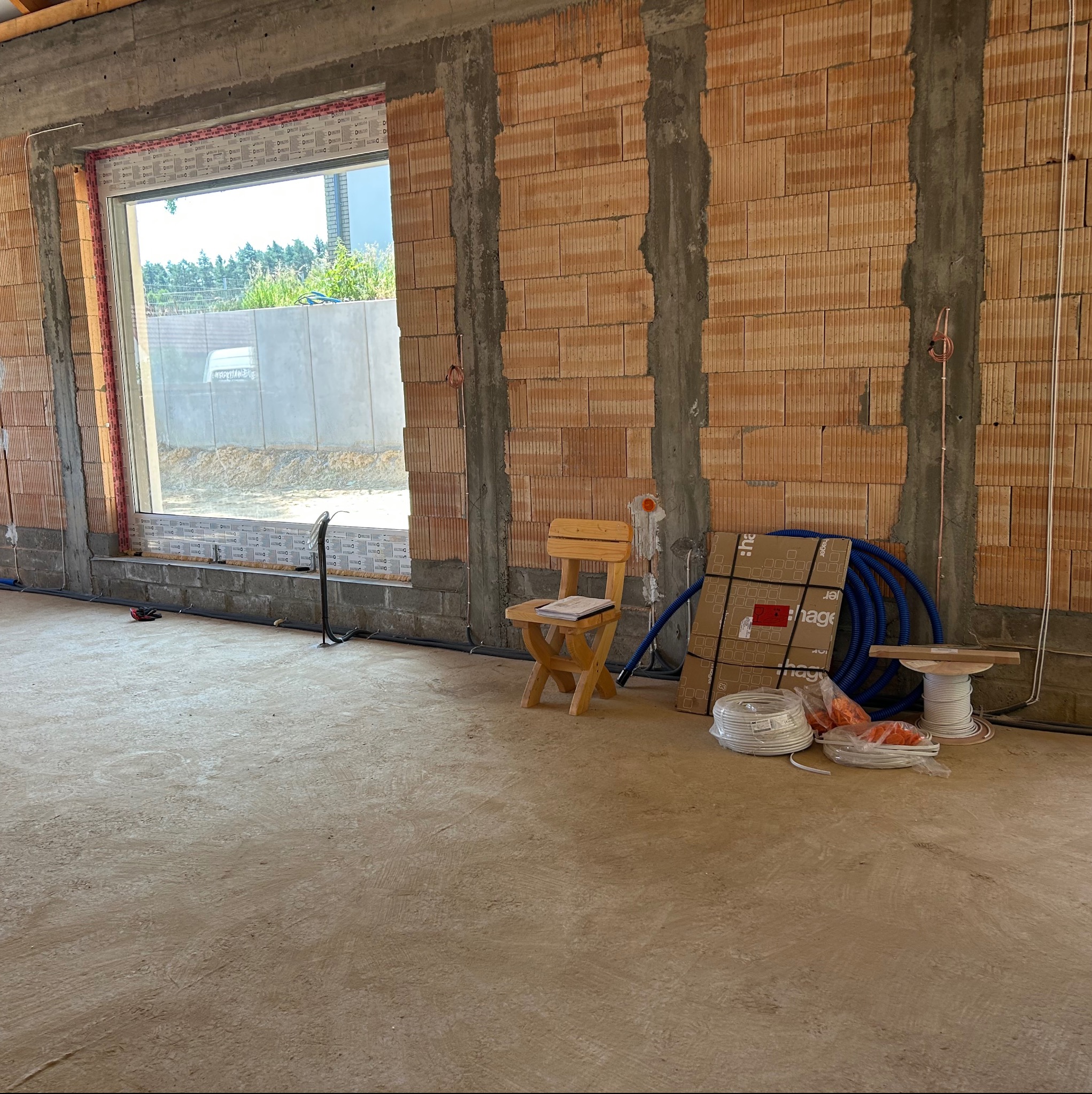 Wnętrze w trakcie budowy z widocznymi przewodami elektrycznymi w ścianach, niebieską rurą instalacyjną i zwojami kabli na podłodze. Widoczne okno i krzesło.