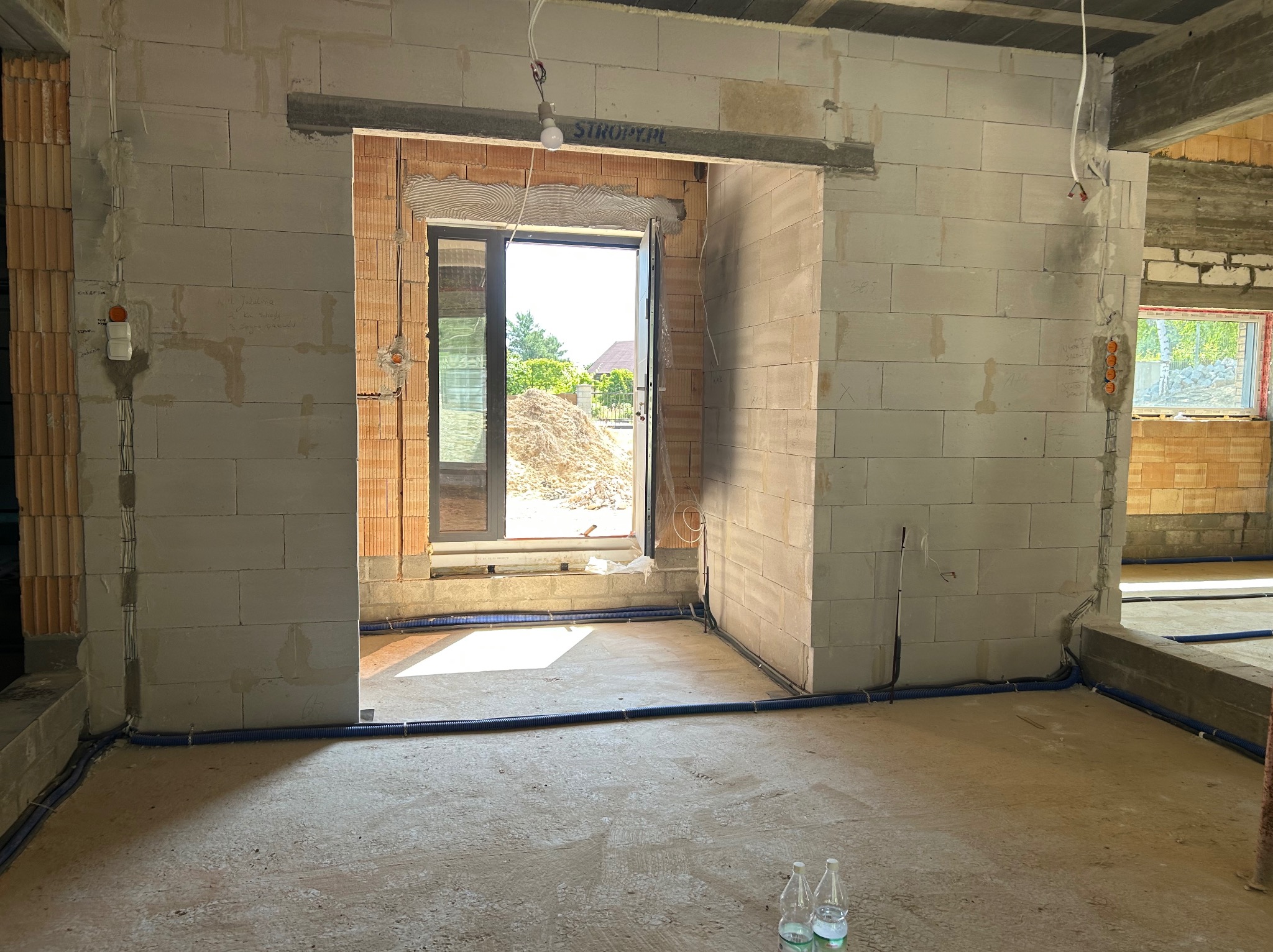 Wnętrze w stanie surowym: instalacja elektryczna w peszlach, ściany z bloczków, widoczne puszki elektryczne. Otwór drzwiowy z widokiem na ogród.