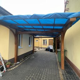 Tomek Cholewinski - Zadaszenie drewniane z niebieską plandeką nad podjazdem z kostki brukowej, widoczne elementy konstrukcyjne z ciemnego drewna, żółte ściany budynków po bokach, drabina oparta o ścianę po lewej.