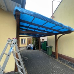 Tomek Cholewinski - Drewniana konstrukcja z niebieskim, przezroczystym zadaszeniem, prawdopodobnie w trakcie montażu lub zabezpieczona przed warunkami atmosferycznymi, widoczna drabina Krause oraz kostka brukowa.