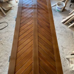 Lakierowane, drewniane drzwi z ukośnym wzorem desek, widoczne w warsztacie stolarskim z resztkami drewna na podłodze.