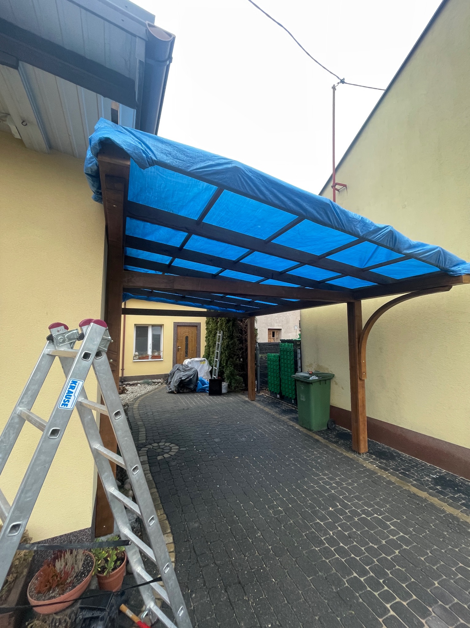 Drewniana konstrukcja z niebieskim, przezroczystym zadaszeniem, prawdopodobnie w trakcie montażu lub zabezpieczona przed warunkami atmosferycznymi, widoczna drabina Krause oraz kostka brukowa.