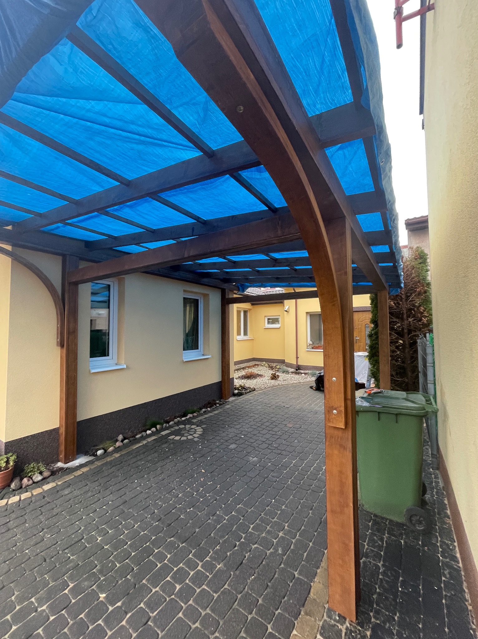 Drewniana konstrukcja z niebieskim zadaszeniem, prawdopodobnie w trakcie budowy w Końskich, z widocznym koszem na śmieci i kostką brukową.