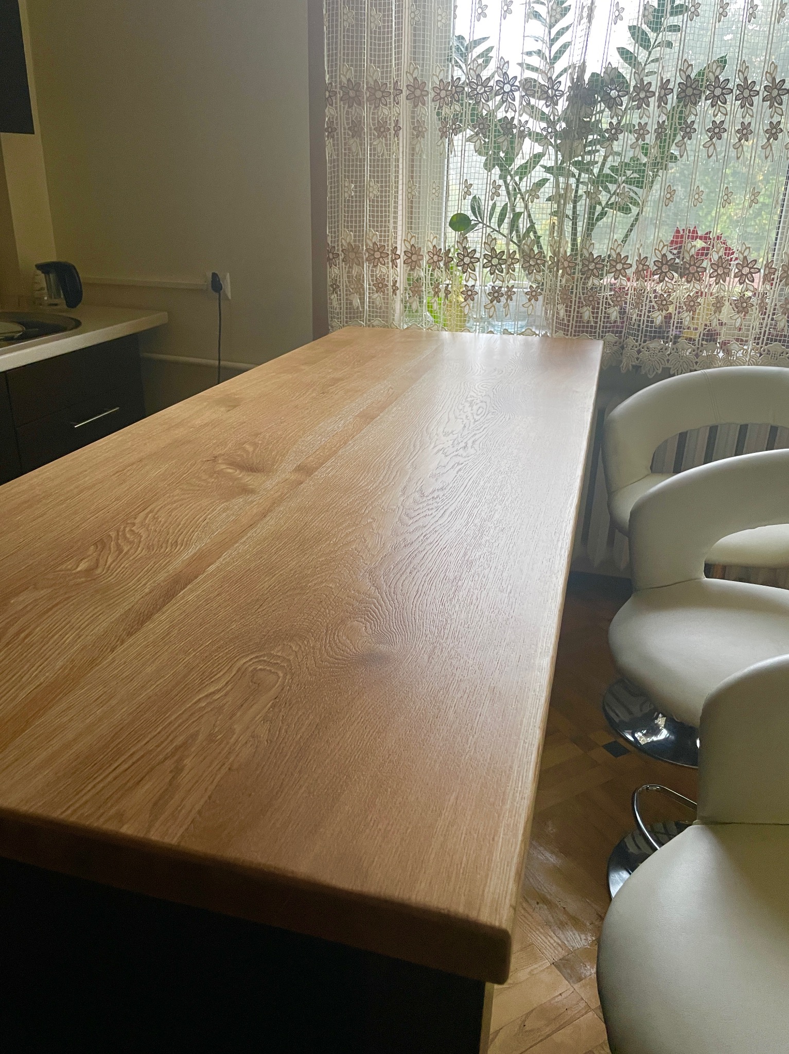 Blat z jasnego drewna z widocznym usłojeniem, stanowiący część wyspy kuchennej, z trzema białymi krzesłami barowymi w tle i kwiatami za oknem.