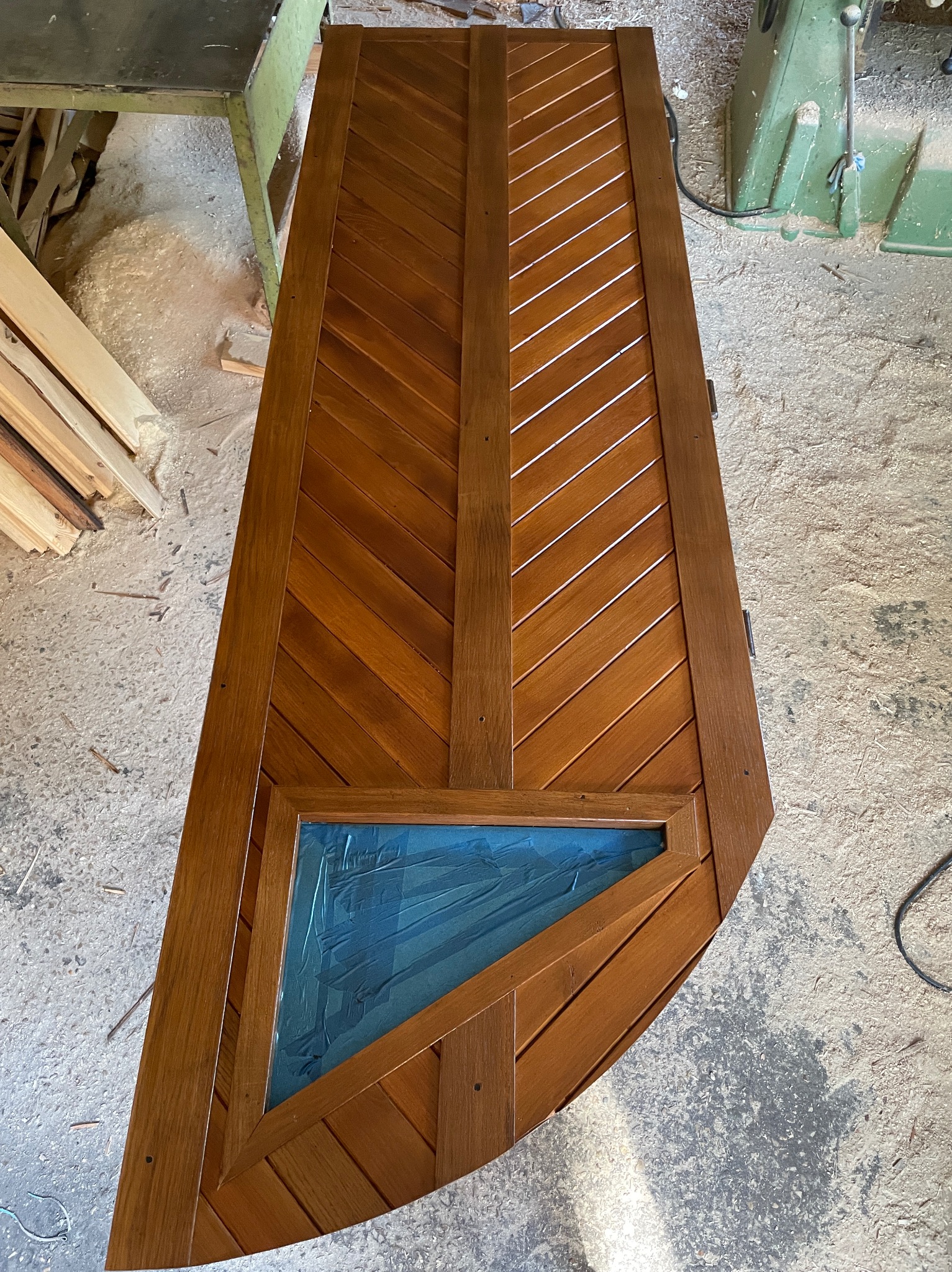 Brązowe drewniane drzwi z ukośnym wzorem i trójkątnym okienkiem zabezpieczonym niebieską folią, leżące w warsztacie stolarskim.