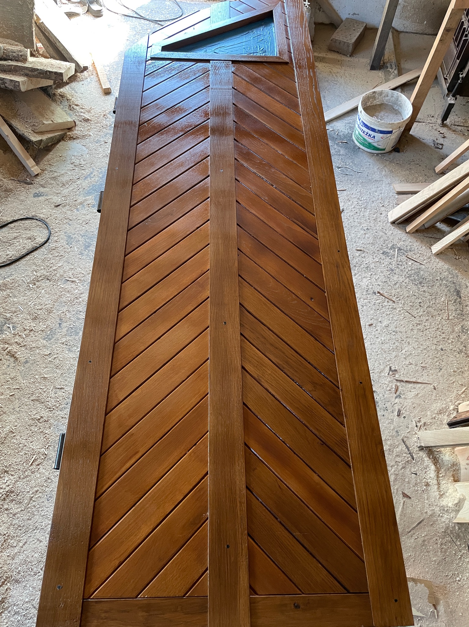 Lakierowane, drewniane drzwi z ukośnym wzorem desek, widoczne w warsztacie stolarskim z resztkami drewna na podłodze.