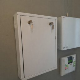 RAFAŁ PRZYBYLSKI WORLD-TEL - Biała szafka z kluczykami, obok centrala alarmowa i panel sterowania. Elementy systemu zabezpieczeń zamontowane na ścianie.