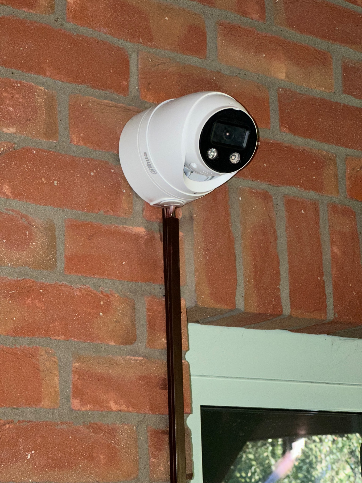 Biała kamera monitoringu marki Dahua zamontowana na czerwonej ceglanej ścianie obok okna z zieloną ramą. Kabel kamery ukryty w brązowej listwie.