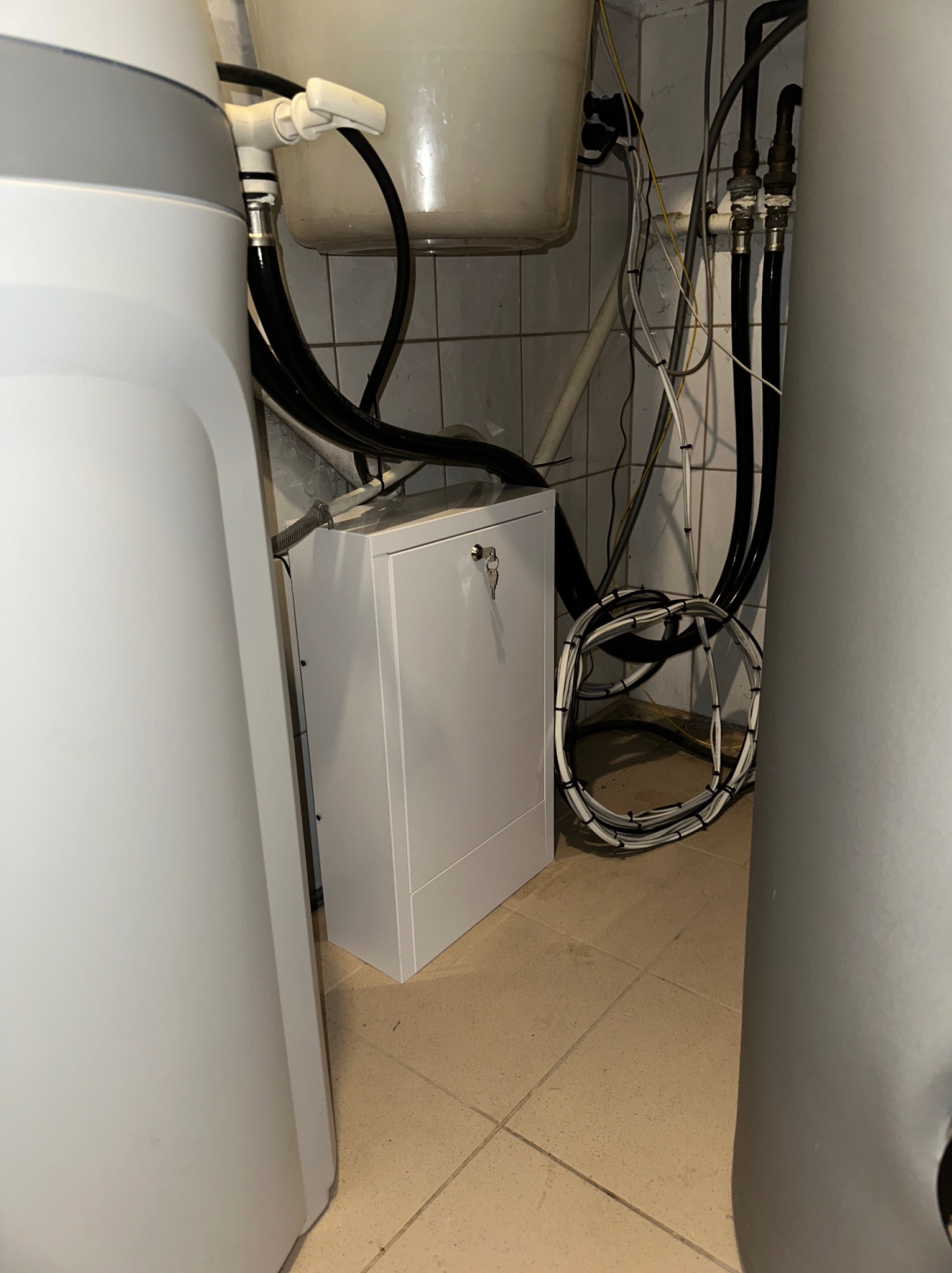 Węzeł wodociągowy z rurami, przewodami elektrycznymi i szafką zabezpieczeń w pomieszczeniu z kafelkami.