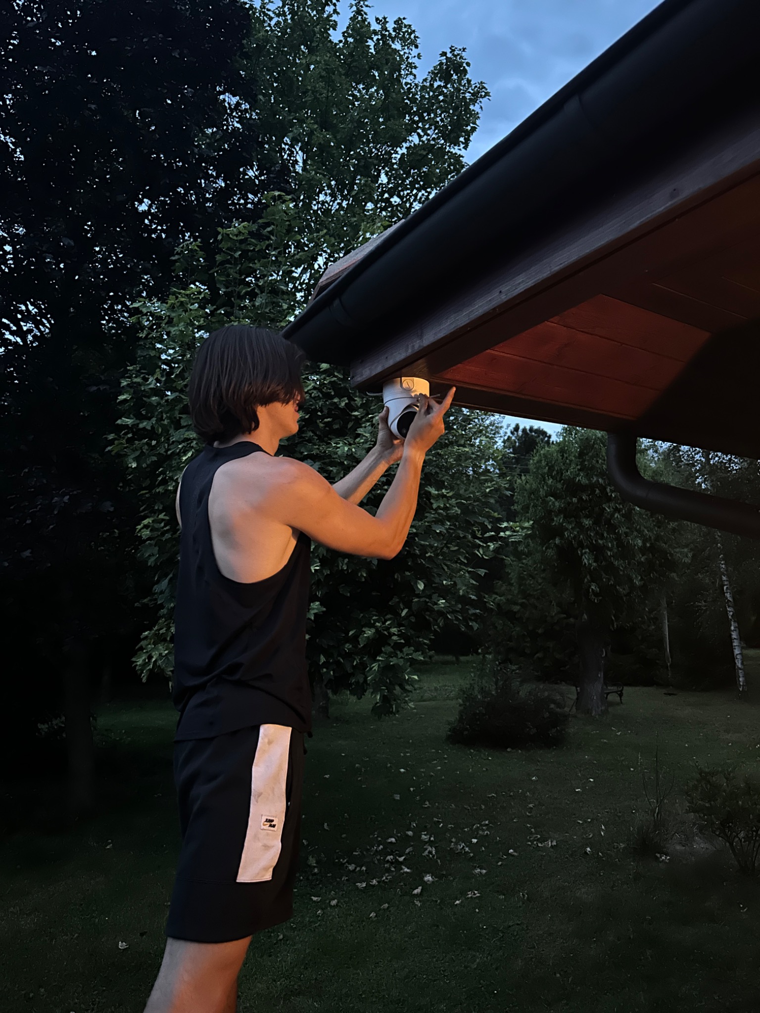 Instalacja kamery monitoringu pod okapem domu, widok na zielony ogród w tle, mężczyzna w czarnej koszulce i sportowych spodenkach montuje kamerę.