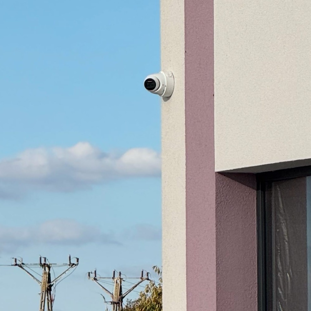 Kamera monitoringu na rogu budynku z kremową elewacją i fioletowym akcentem, na tle błękitnego nieba z chmurami i słupów energetycznych.